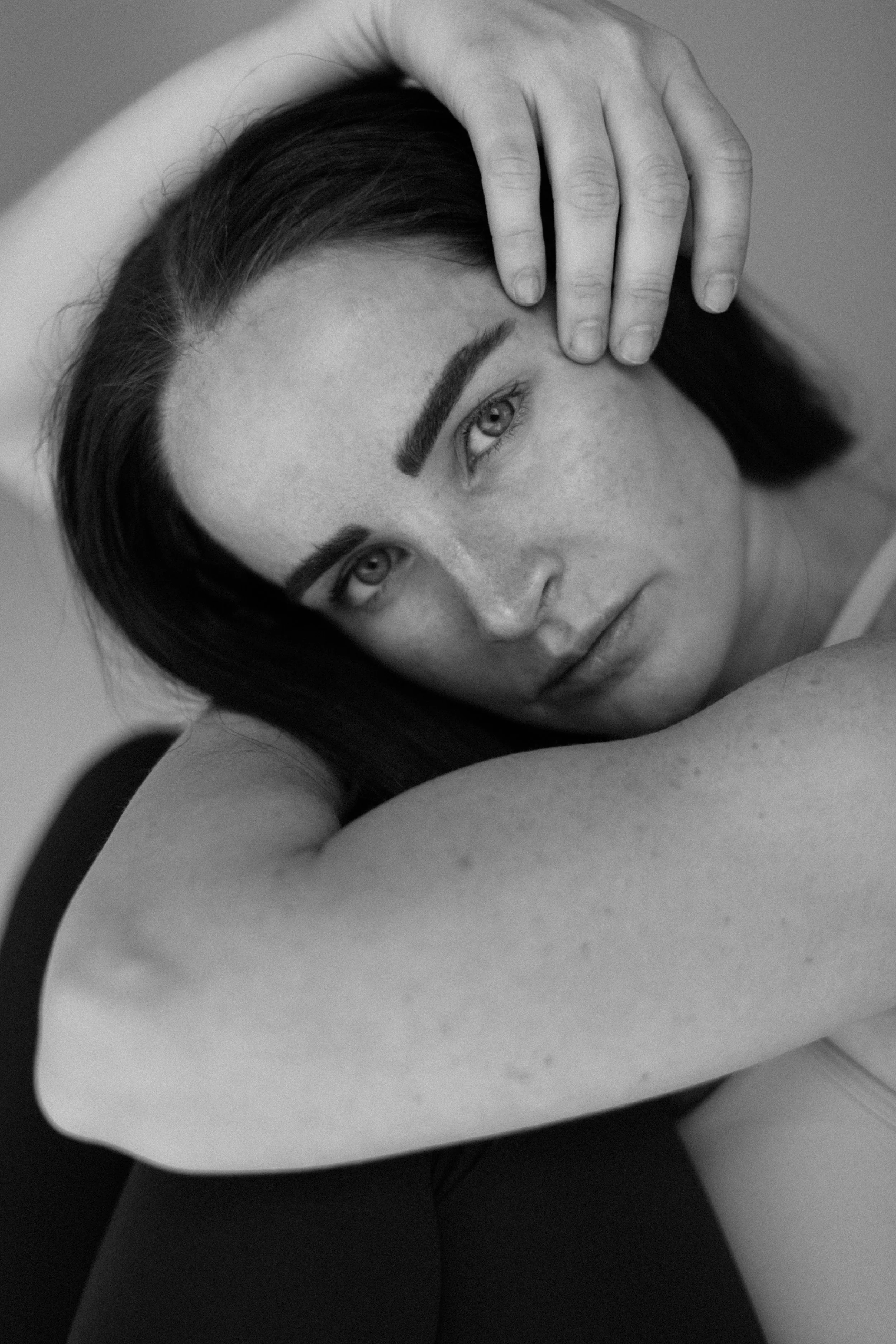 A black and white close-up portrait of a woman with dark hair, intense eyes, and freckles, resting her head on her crossed arms, with one hand touching her forehead.