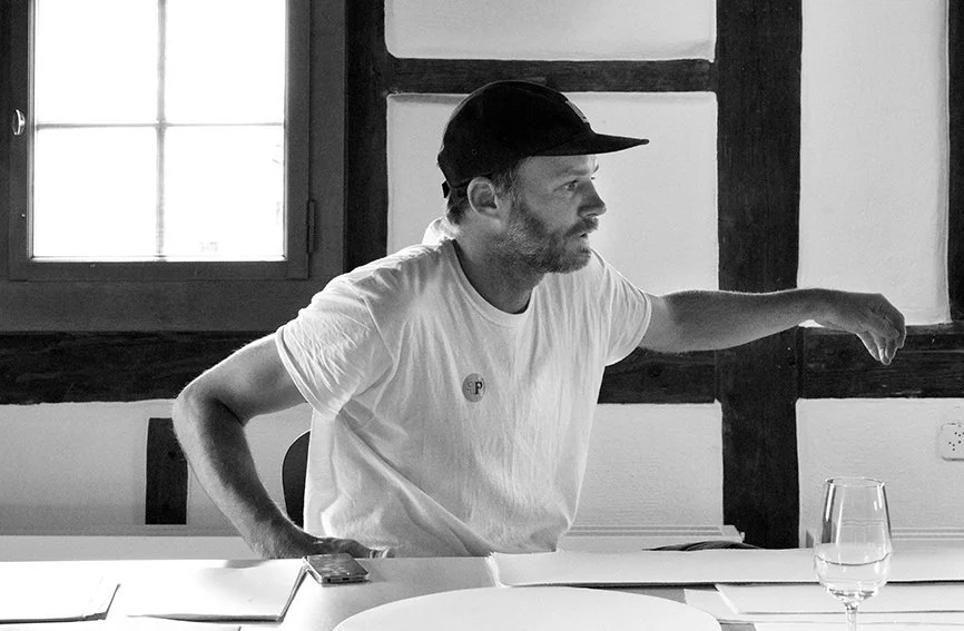 A man sitting at a table in front of a window with wooden framing, wearing a baseball cap and white T-shirt, with his right arm extended across the table.