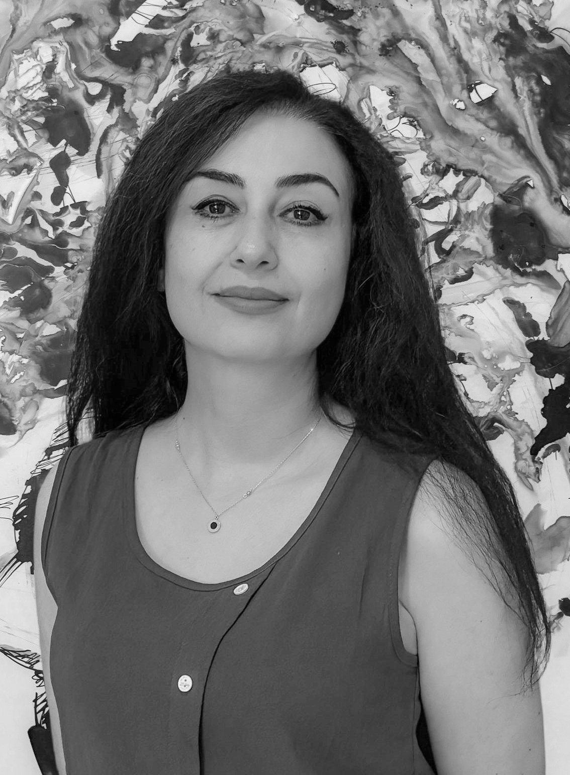 A woman with long dark hair, makeup, and a jewelry necklace, smiling slightly, standing in front of an abstract black and white background.