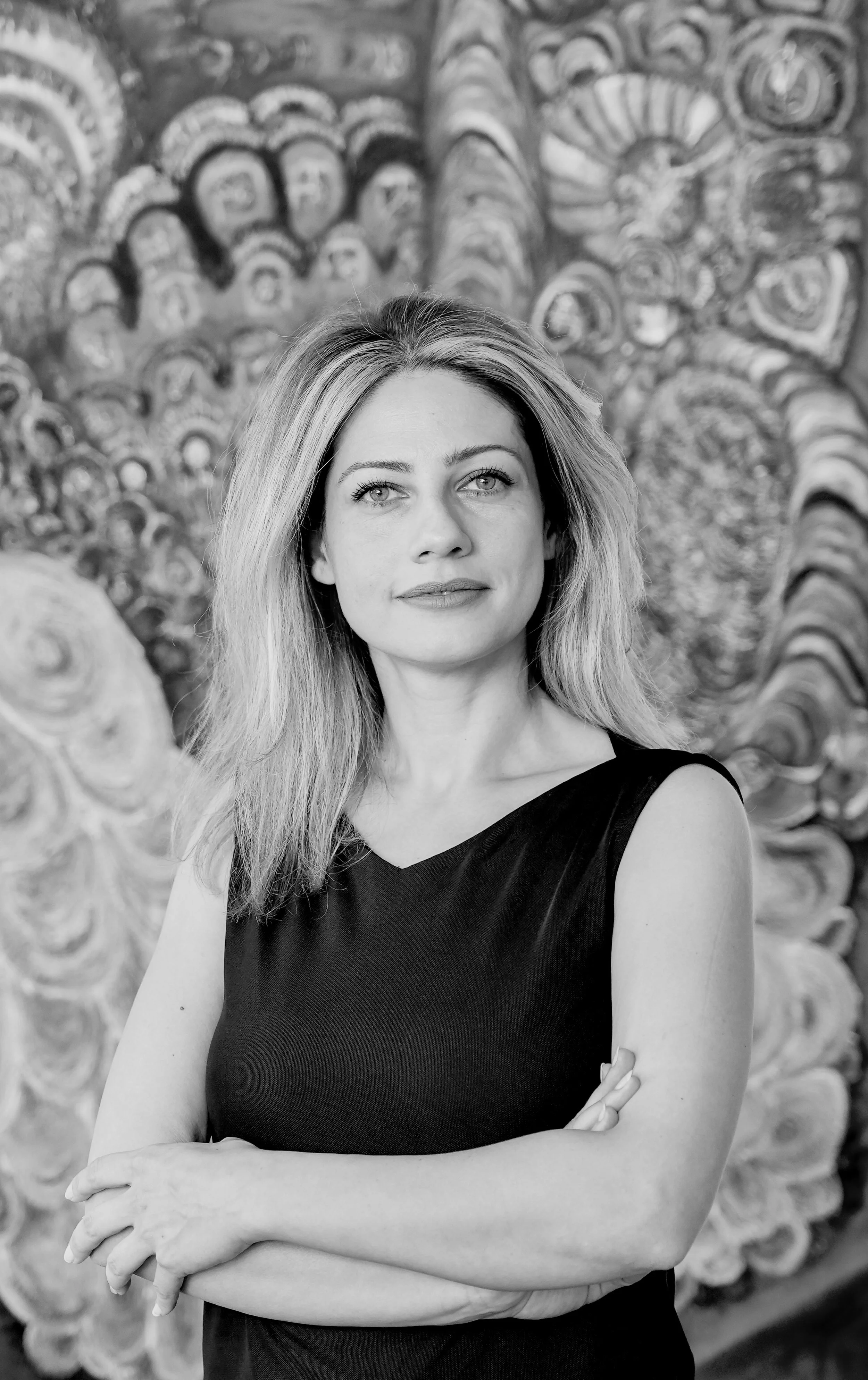 Black and white photo of a woman with light-colored, wavy hair standing in front of an abstract, swirling painted background. She is wearing a sleeveless black top with her arms crossed and looking slightly upwards.