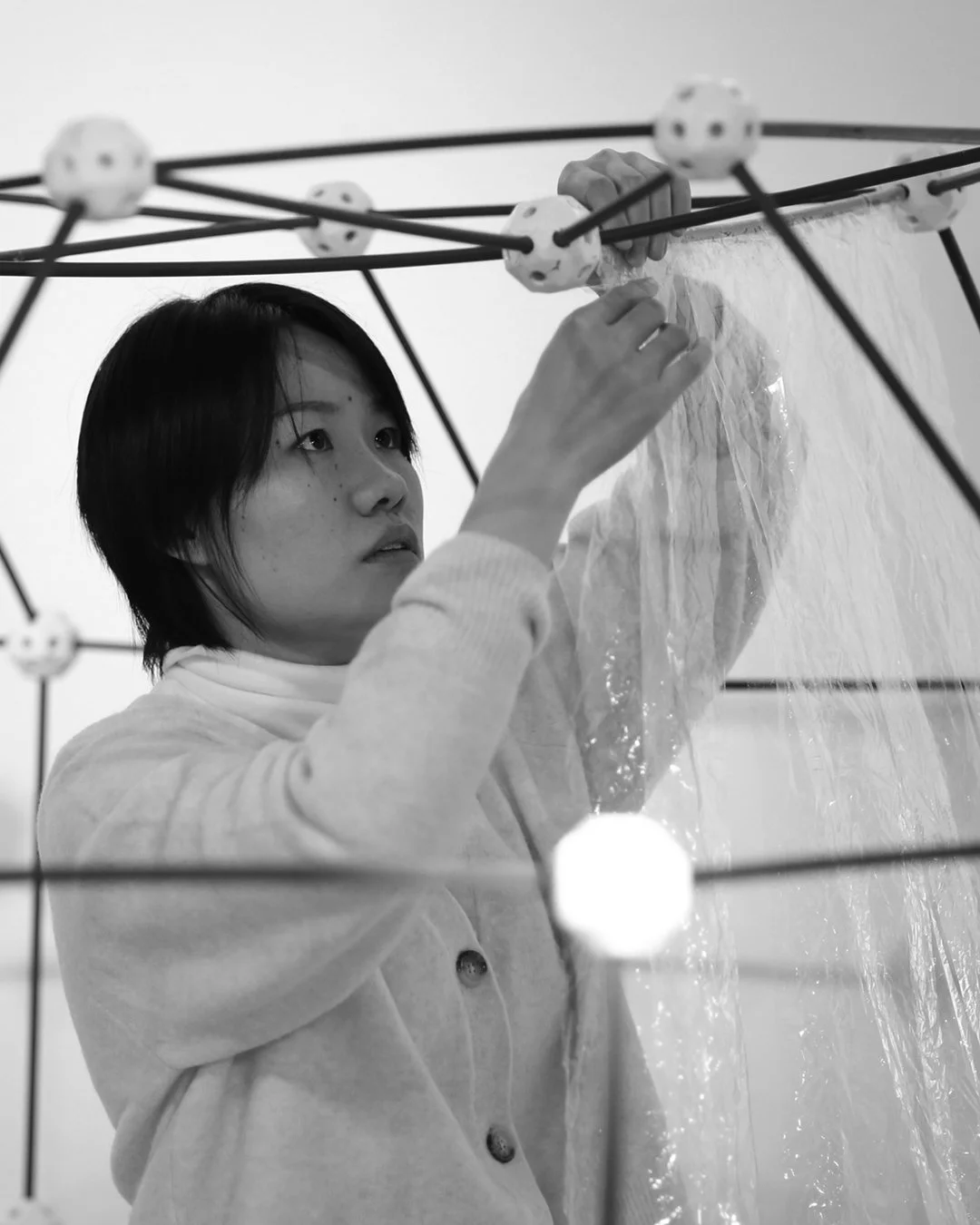A woman with short black hair and a light-colored sweater working inside a spherical structure with interconnected rods and small white spherical nodes, adjusting or assembling a plastic sheet.