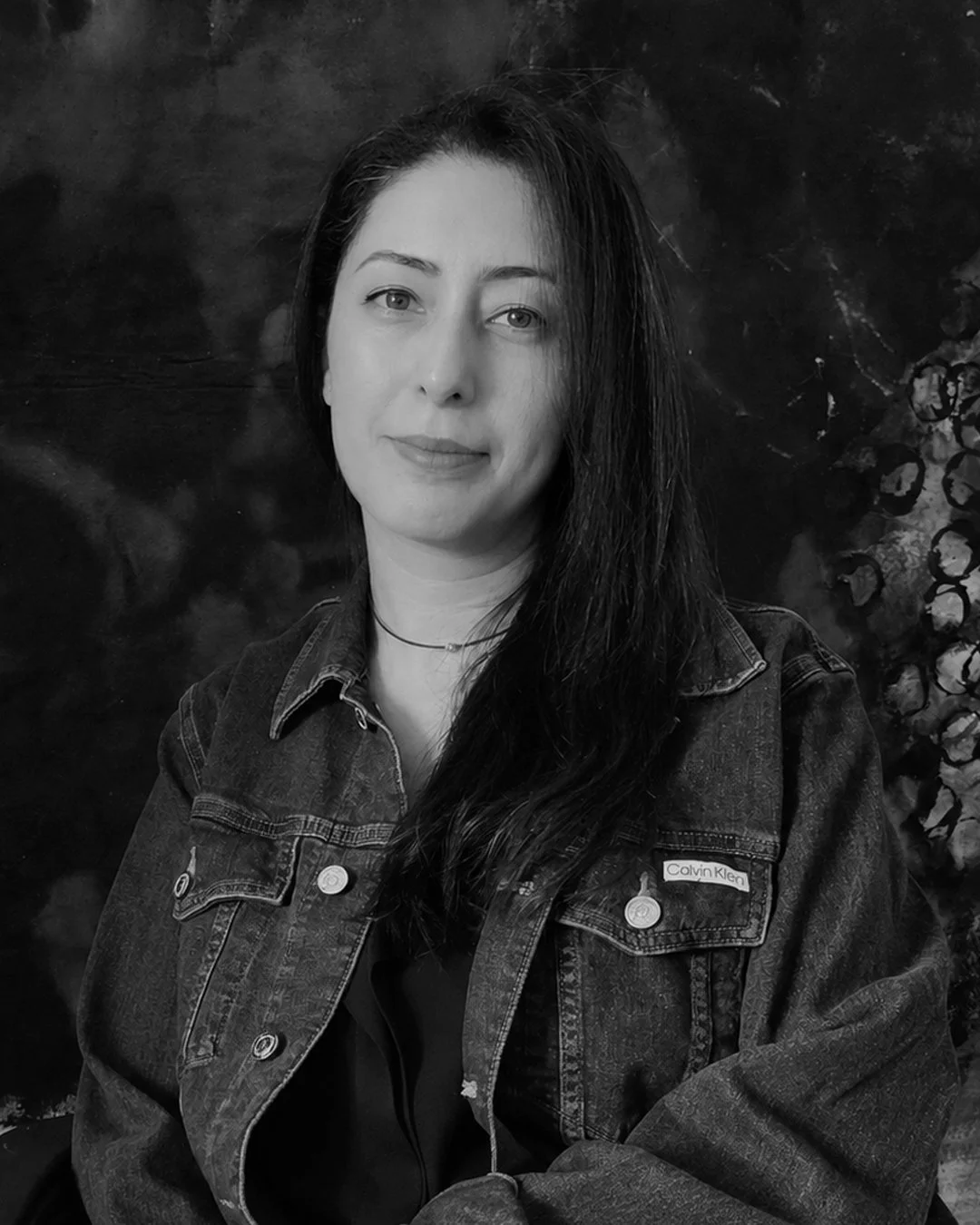 Black and white portrait of a woman with long, dark hair wearing a denim jacket and a choker necklace, standing in front of a textured dark background.