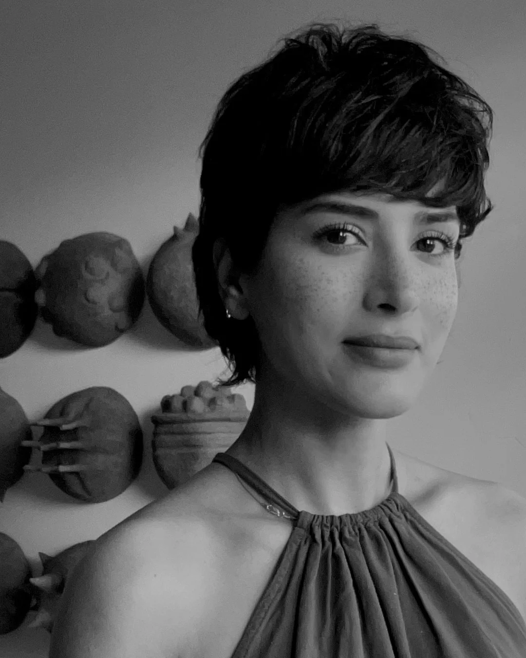 Black and white portrait of a woman with short, wavy hair, standing in front of a wall decorated with artichoke-shaped objects.