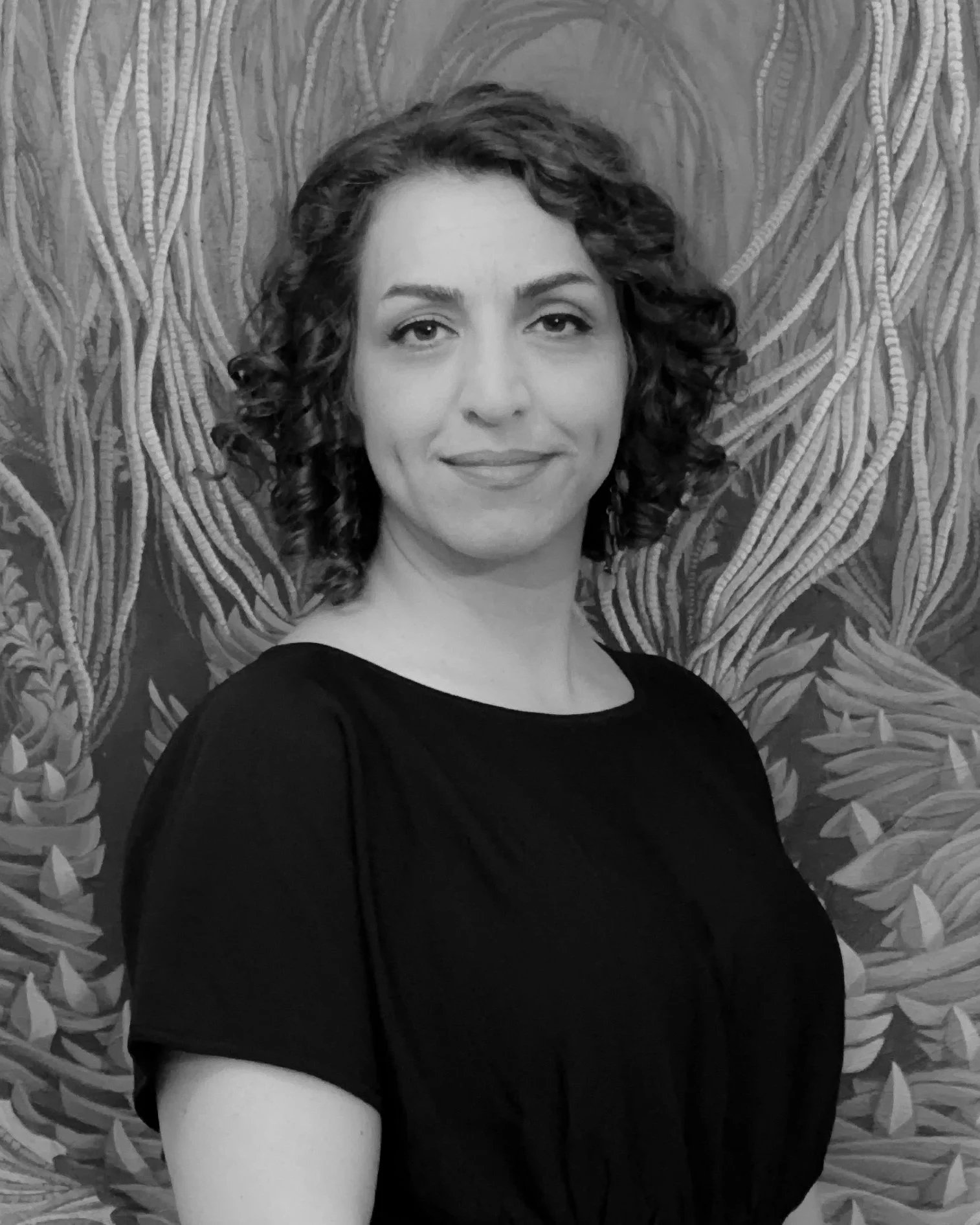 A woman with shoulder-length curly hair, wearing a black top, is smiling gently at the camera. The background features a detailed, leafy patterned wallpaper.
