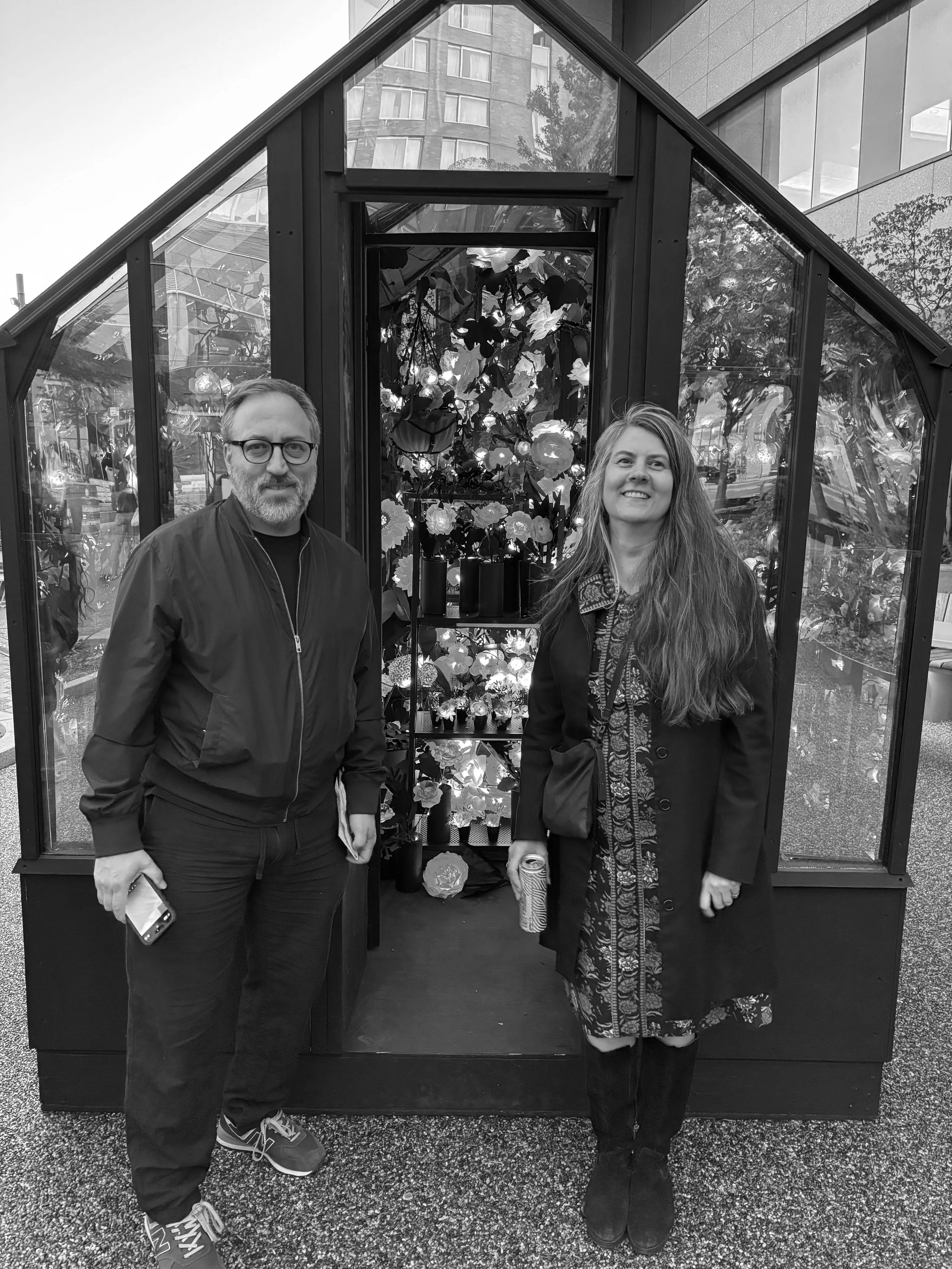 A man and woman standing in front of a greenhouse filled with flowers, smiling at the camera.