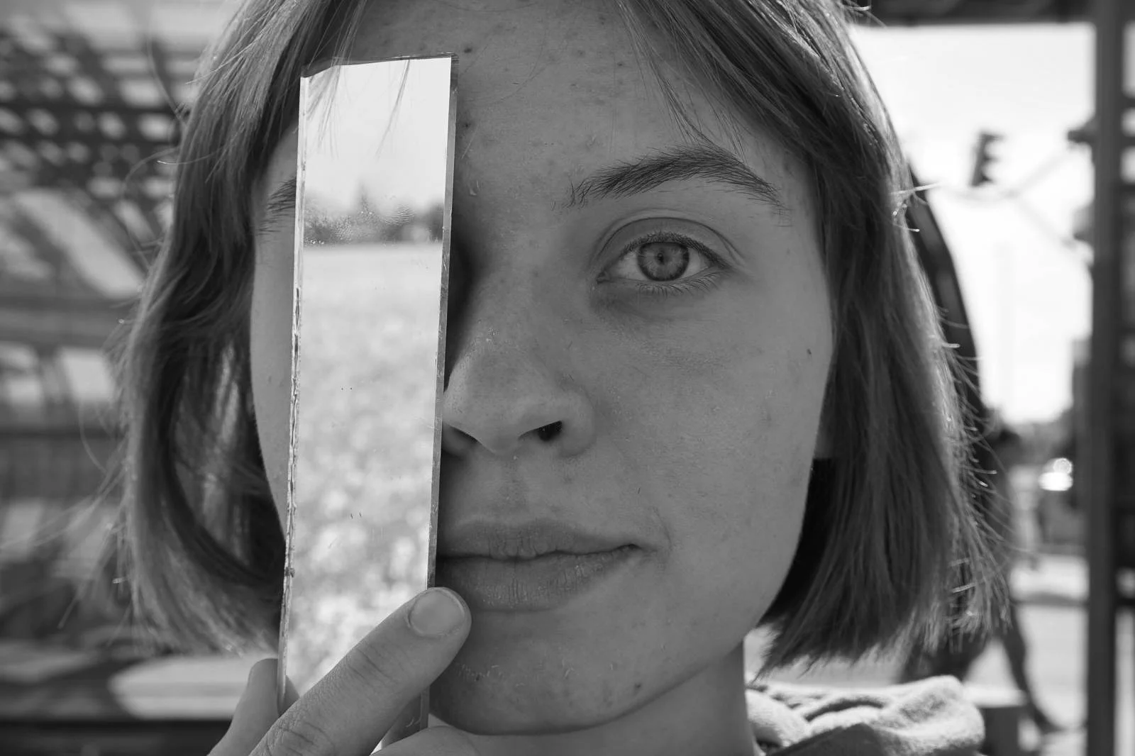 A woman with short hair holds a mirror reflecting her face, covering part of her face with her finger, outdoors on a sunny day.