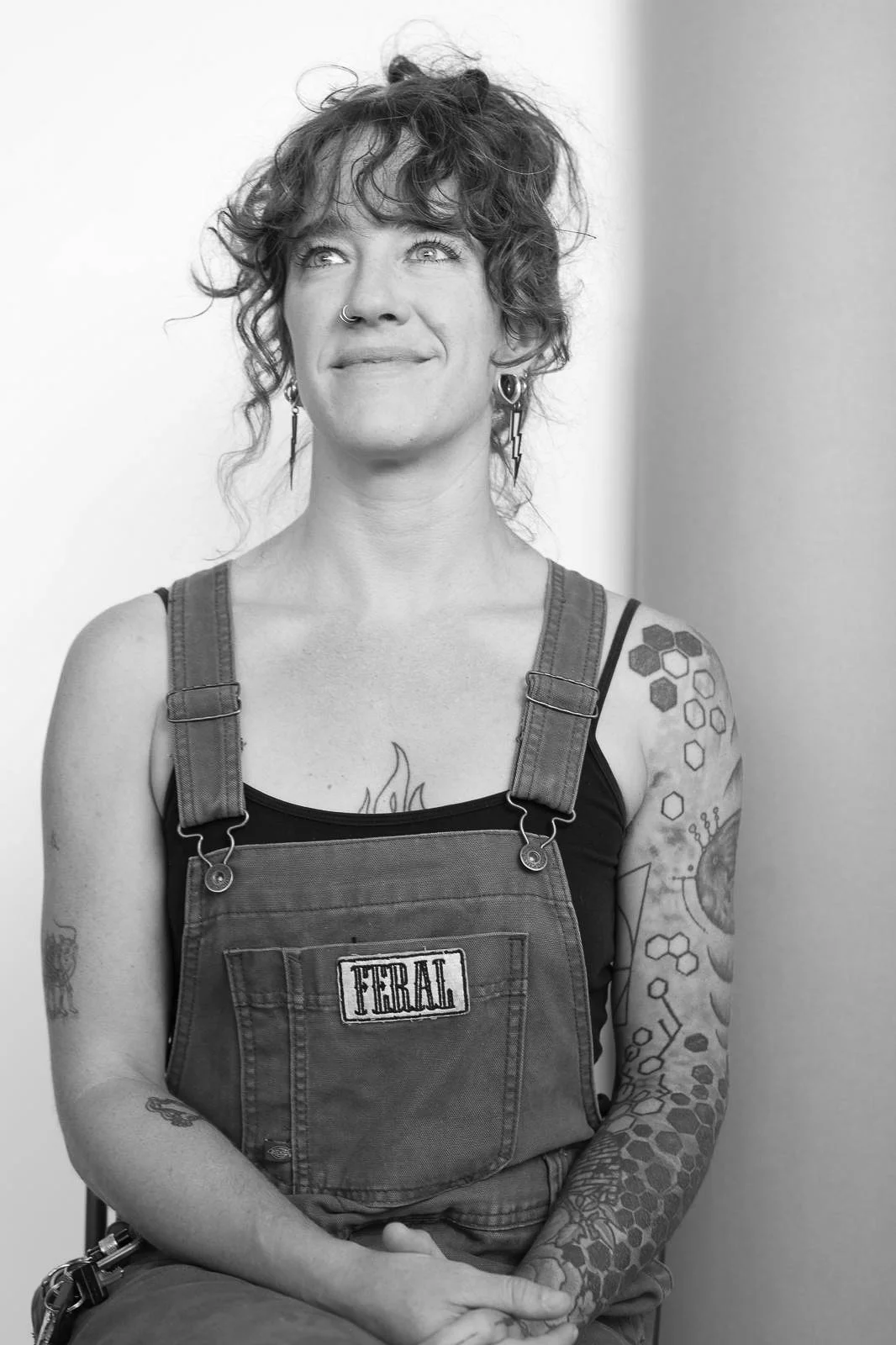 Black and white photo of a smiling woman with curly hair, tattoos, and a nose piercing, wearing overalls labeled 'Feral' and a tank top.