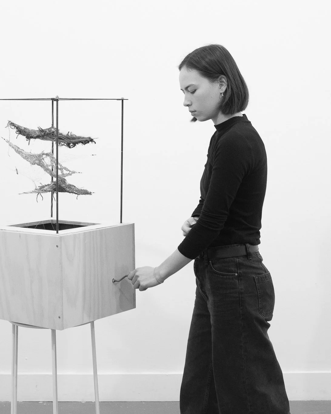 A woman with short hair wearing a black top and dark jeans operating a wooden machine with a handle in a gallery or exhibition space, with art pieces on display.