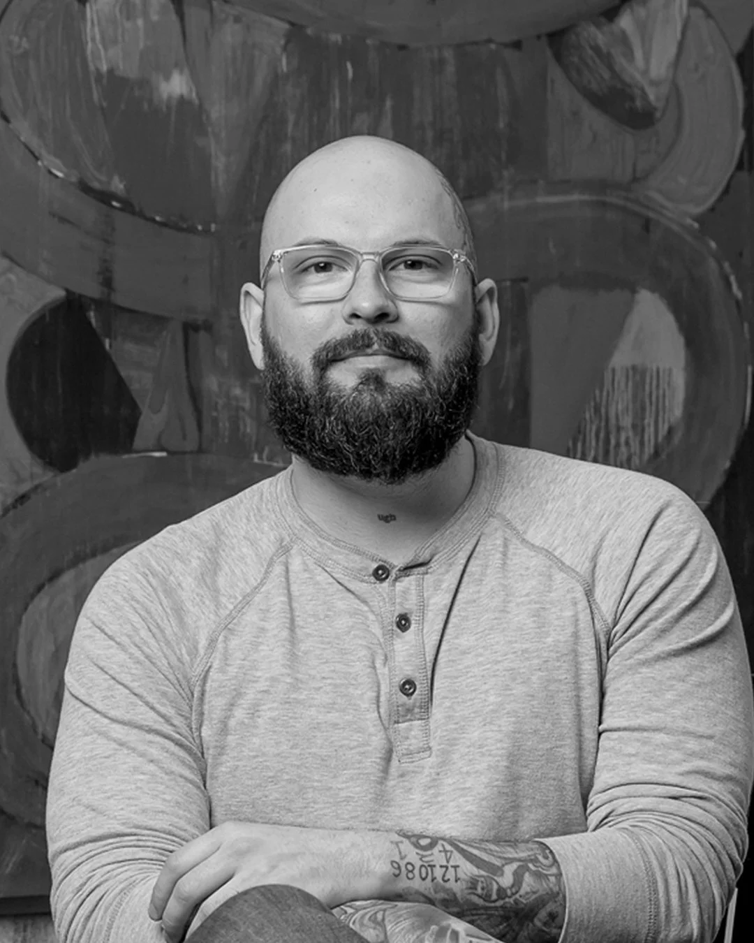 A black and white photo of a bald man with a beard wearing glasses and a long-sleeve henley shirt, sitting with his arms crossed against a background of textured abstract art.