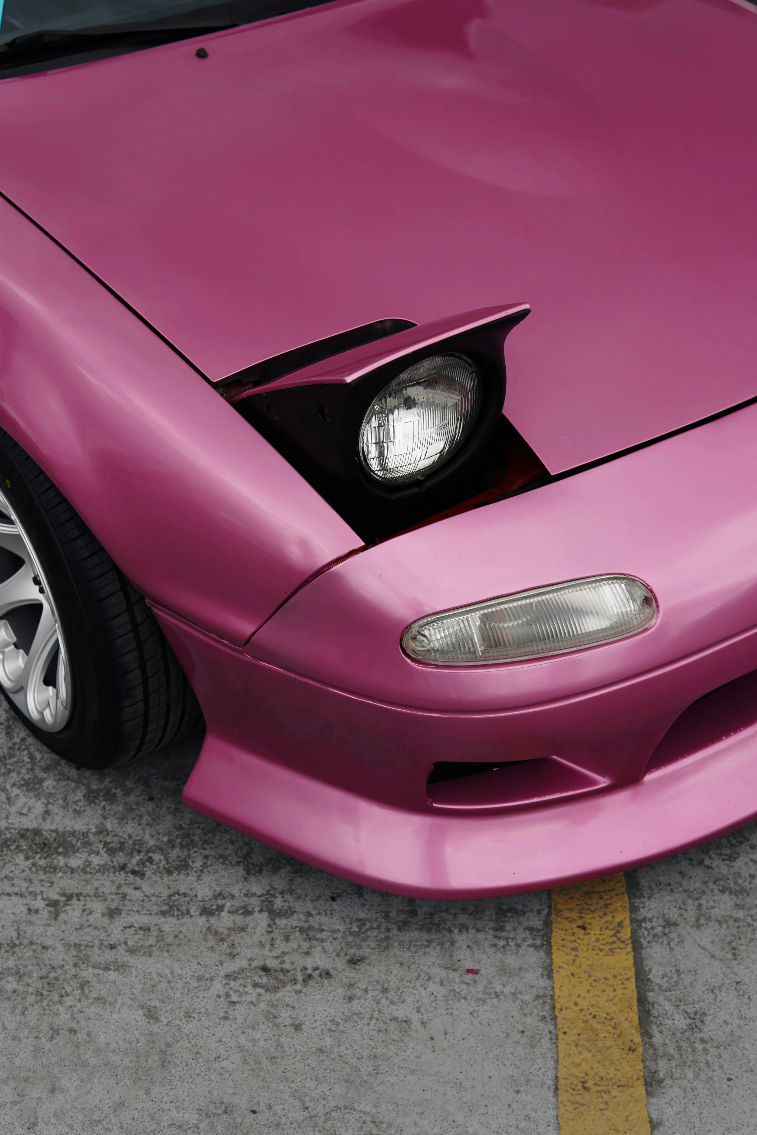 Close-up of a pink sports car front end, featuring a headlight, front bumper, and part of a wheel, parked on a concrete surface with a yellow parking line.
