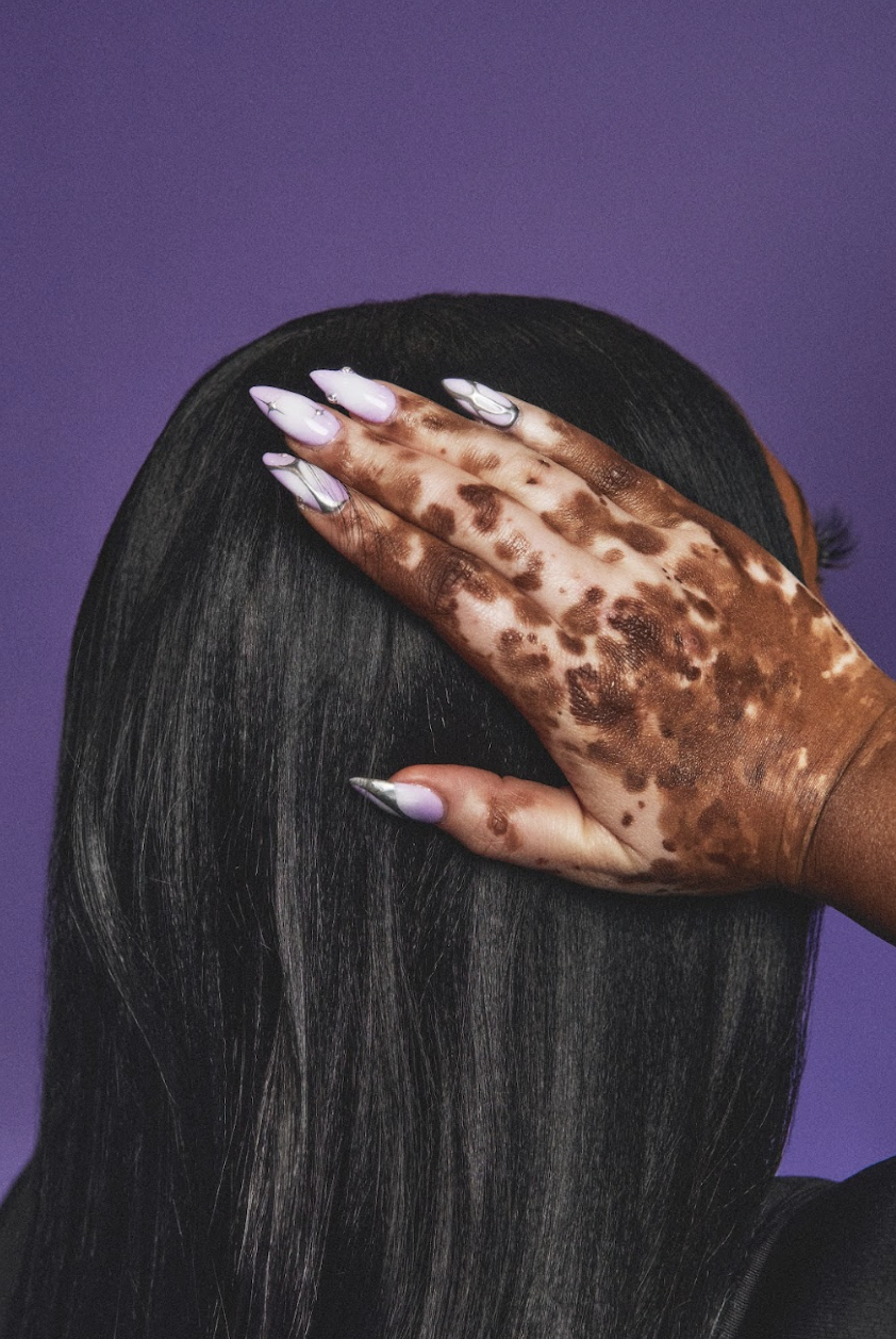 A person with long, straight black hair covering their face, holding their hand with a leopard print pattern over their head, with purple background.