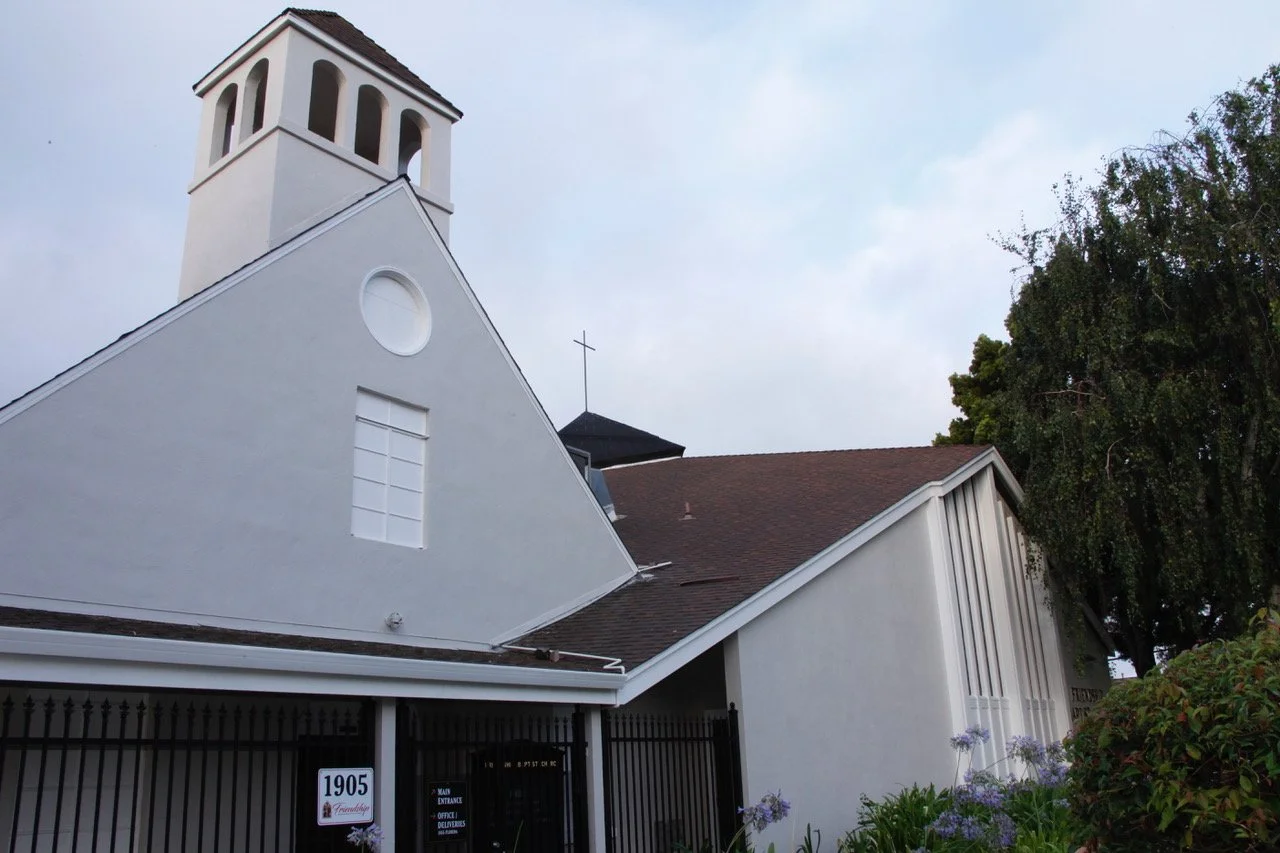 People gathered inside a church, standing at the front near a cross with stained glass windows behind them, during a service or event, with some people sitting in pews and others standing.