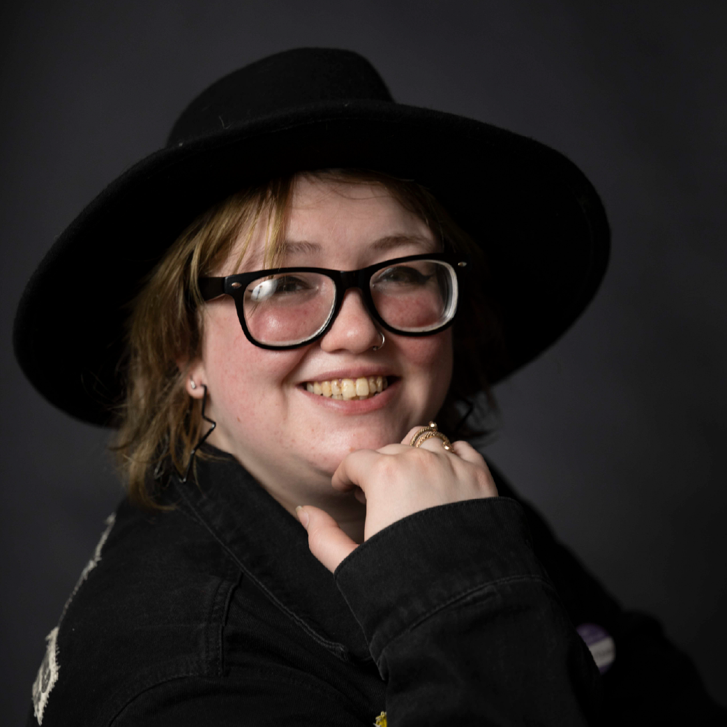 A woman with short, reddish-brown hair, wearing clear glasses, a nose ring, and small earrings, smiling outdoors with green trees in the background. She is wearing a black t-shirt and has a sea turtle tattoo on her left arm.