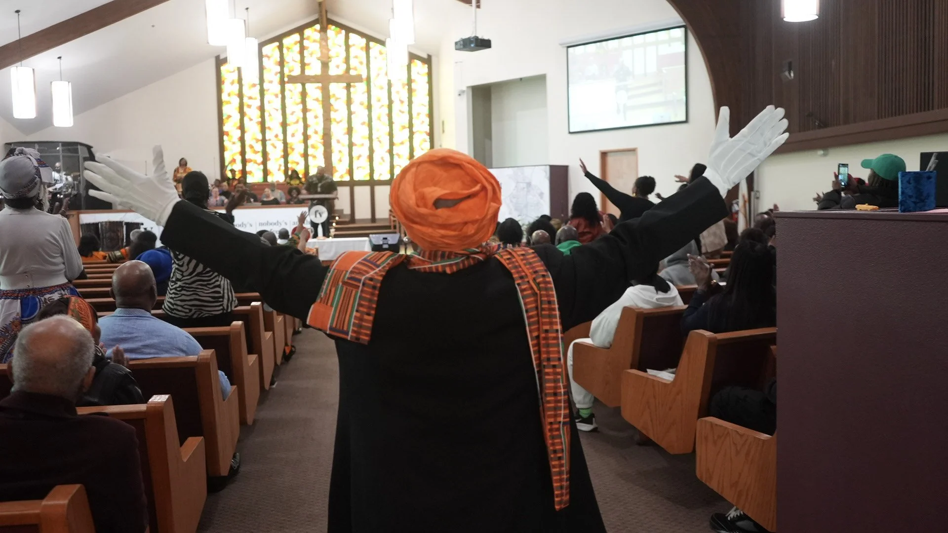 Church congregation stands in wooden pews during worship as a minister in a black robe with an orange headwrap raises their arms toward the front of the sanctuary.