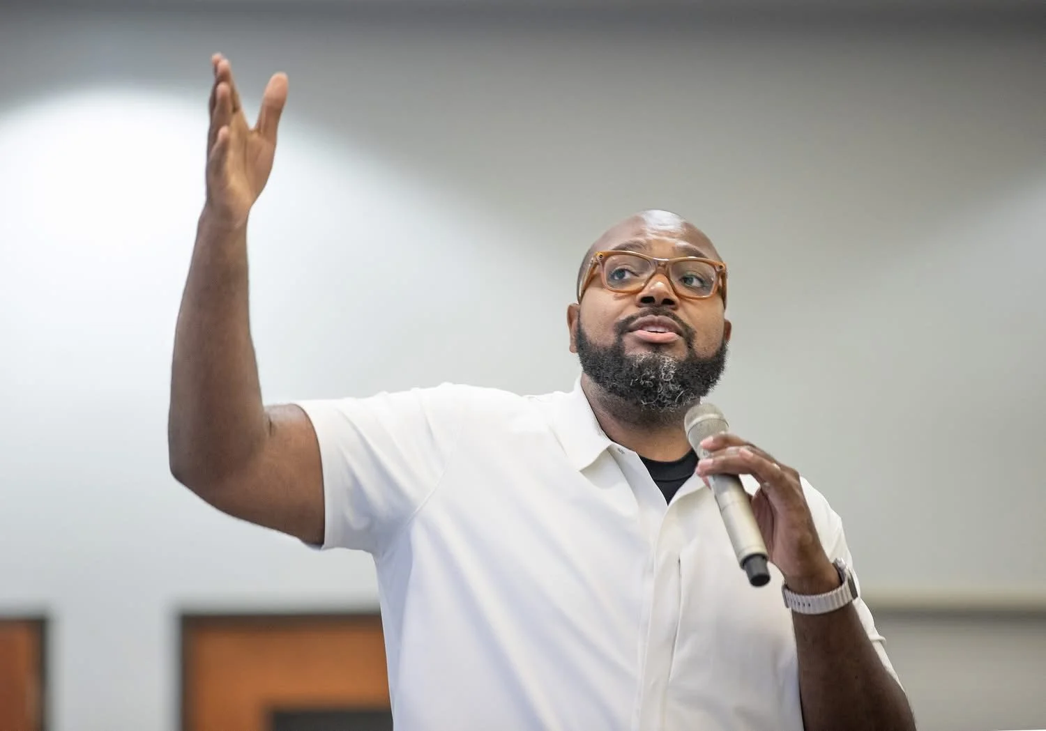 Man wearing glasses and a white polo shirt speaks into a handheld microphone while raising his hand, addressing an audience indoors.