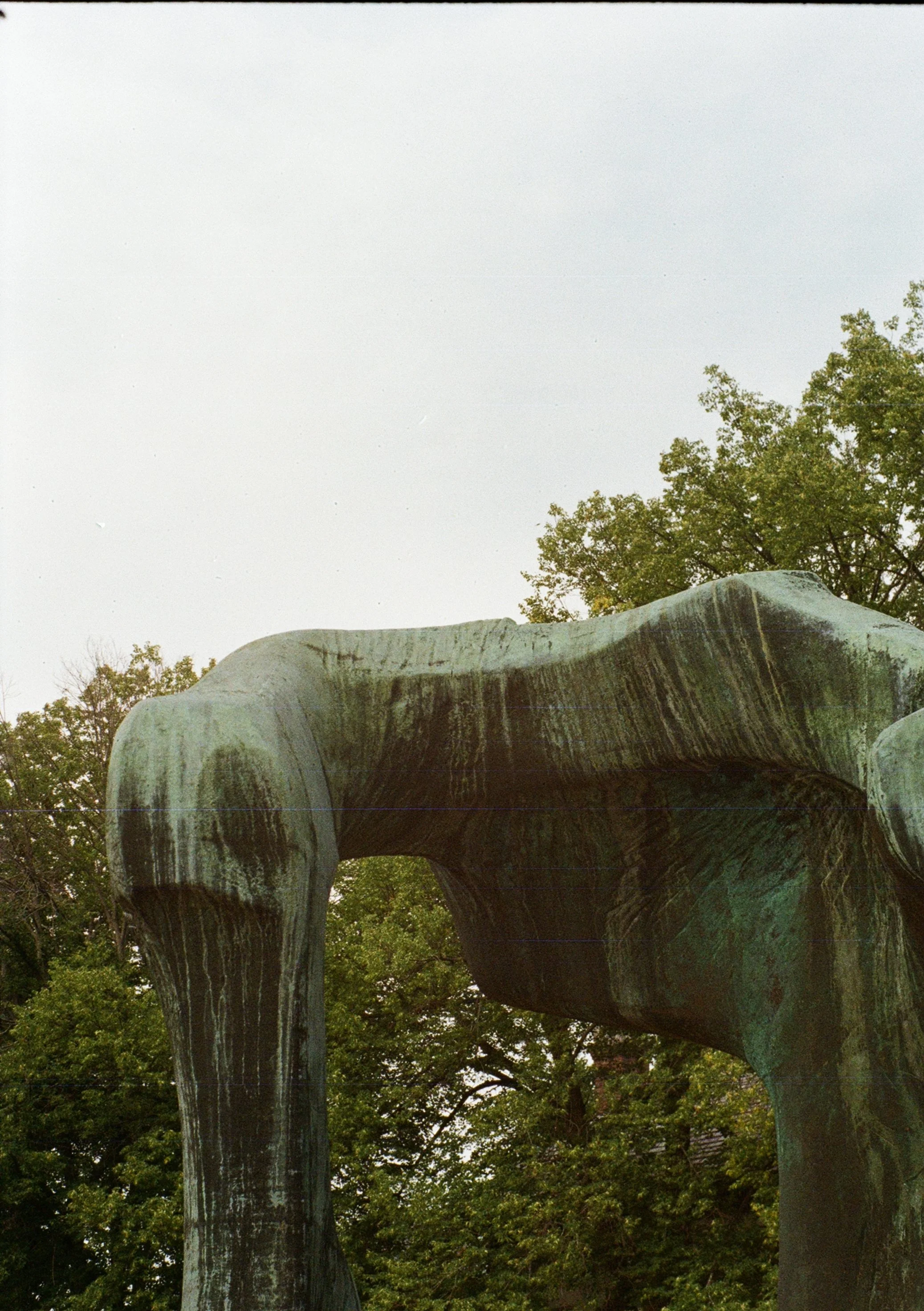 Close-up of a large, weathered bronze sculpture with four legs, set against a background of green trees and a cloudy sky.