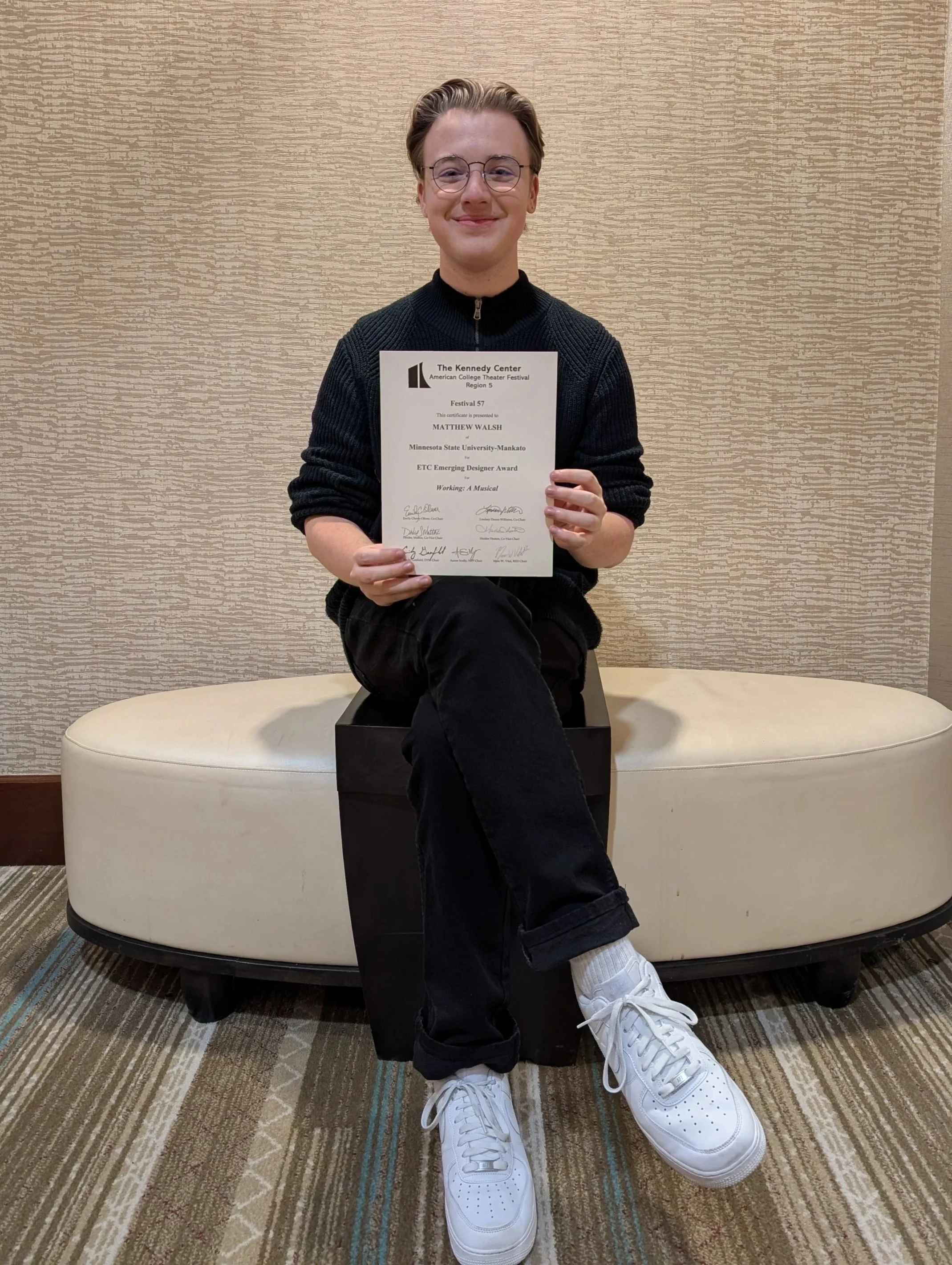 Matthew Bryan Walsh with his signature glasses, wearing a black sweater and pants, sitting on a bench in a room with beige textured walls, holding a certificate for the ETC Emerging Designer Award and smiling at the camera.