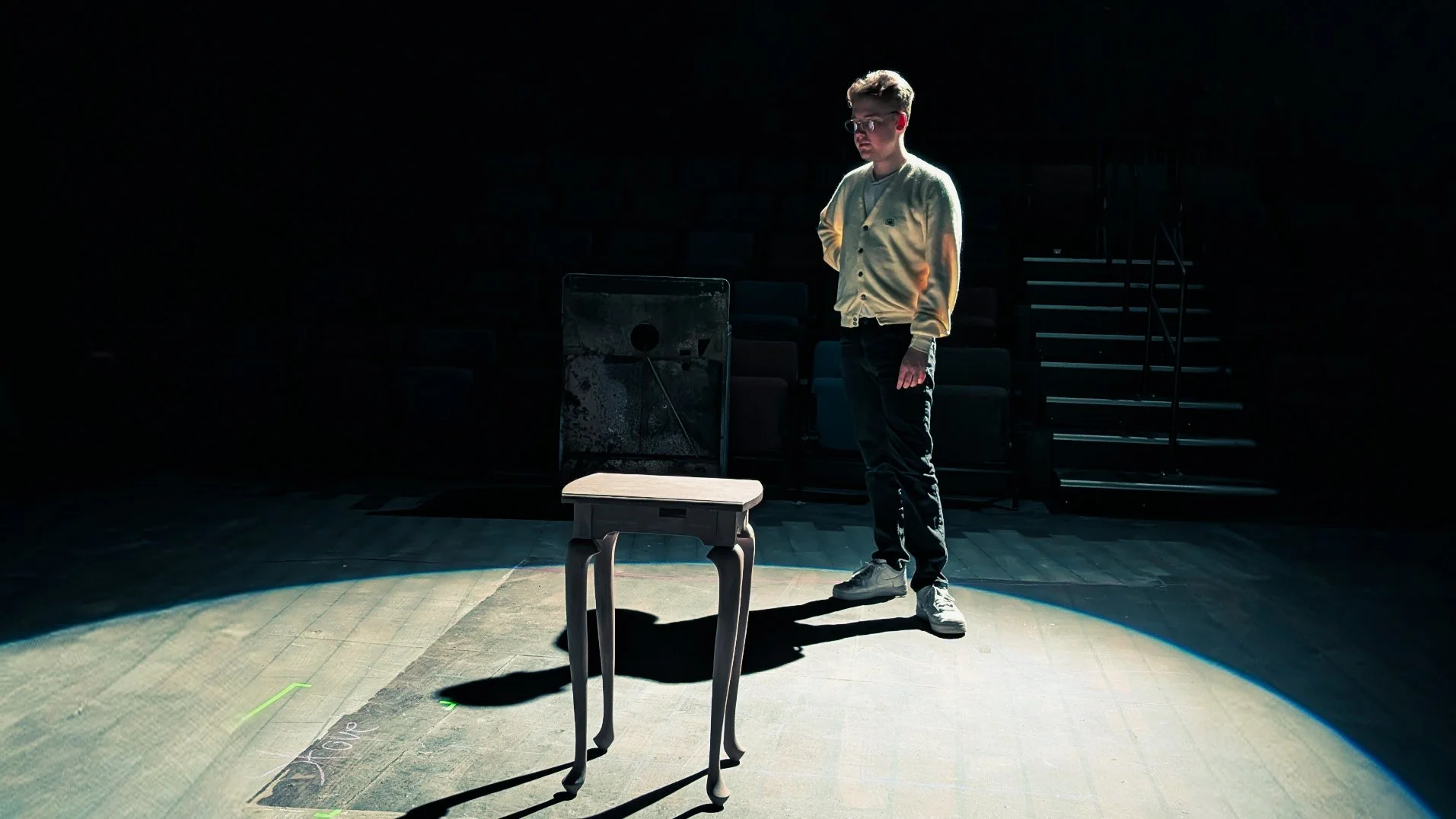 Matthew Bryan Walsh stands in a spotlight on a dark theater stage, facing a small wooden table with curved legs. There are empty seats and stairs in the background. He is in the midst of lighting focus for  The Birds by Conor McPherson.