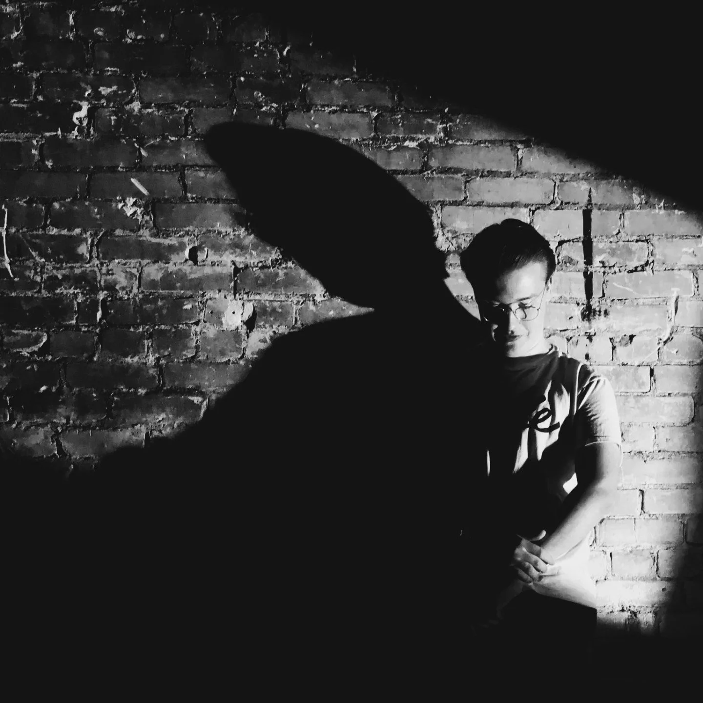 Matthew Bryan Walsh with his signature glasses standing against an old brick wall, with his body partially illuminated and casting a large shadow. Photographed by Jarod-Connor Moss at the Southern Theater in Minneapolis, Minnesota.