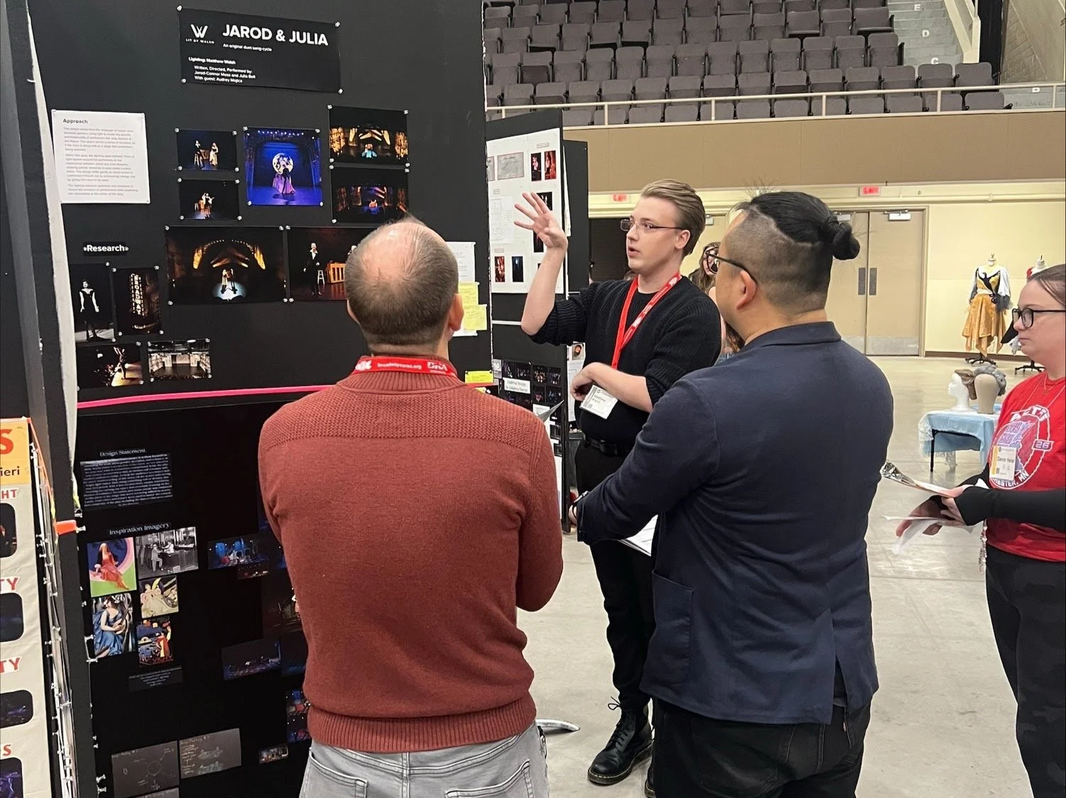 Matthew Bryan Walsh is giving a presentation on a black display board at an indoor event, with three people listening and engaging in the discussion. He is dressed in his signature black outfit with glasses.