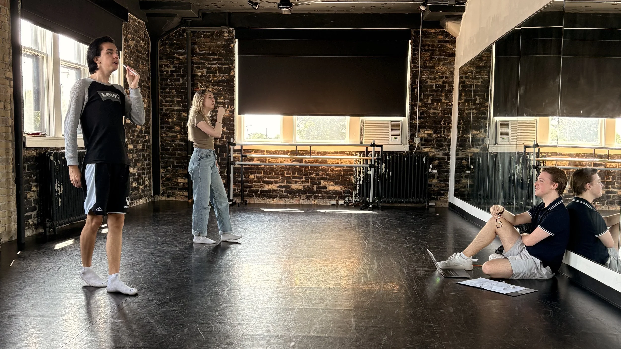 Three young people in a dance studio with exposed brick walls and large mirrors. Matthew Bryan Walsh sitting on the floor next to a laptop, Jarod is standing near the mirror, and Julia is standing near the window. All are casually dressed.