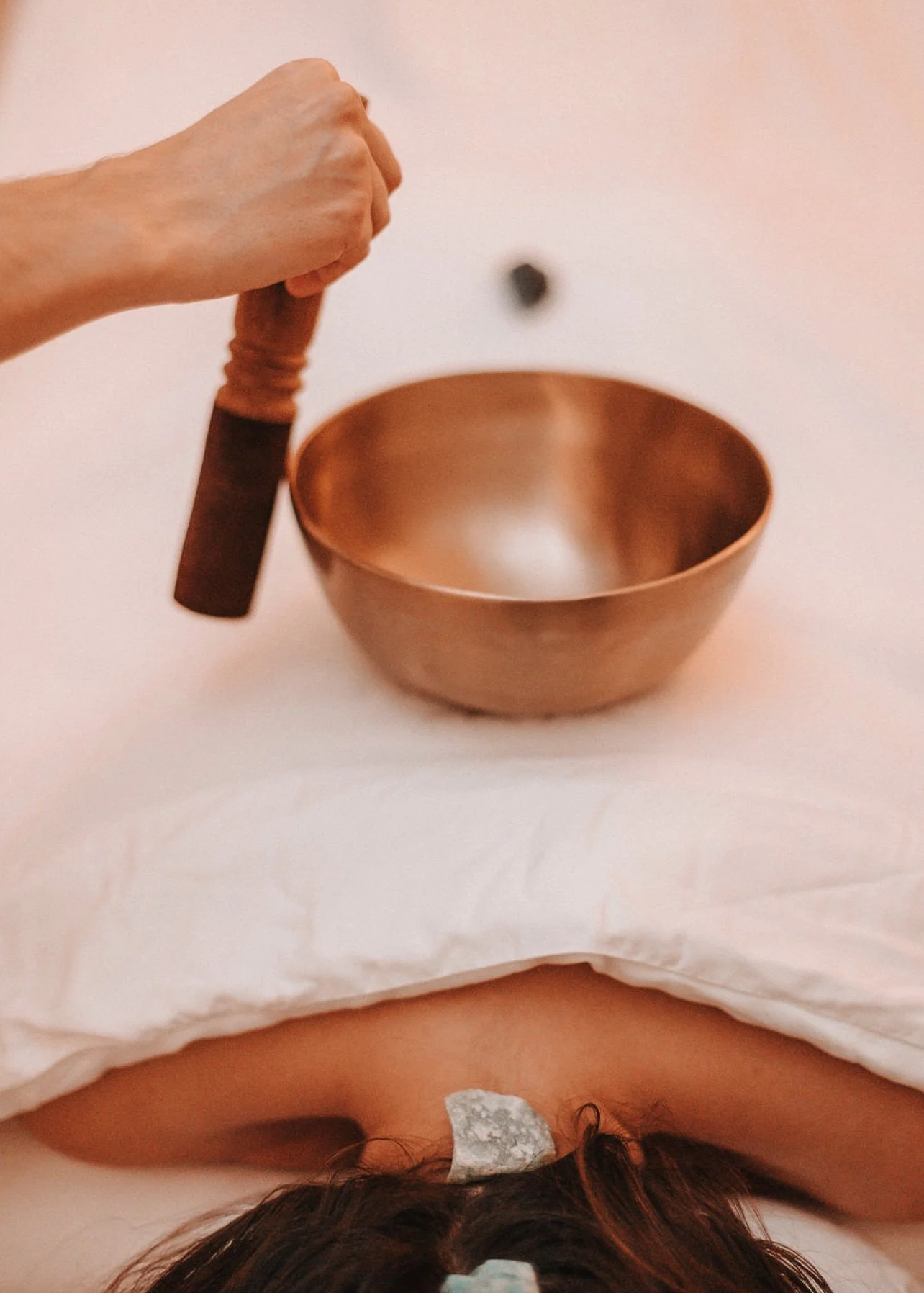 A person is about to use a mallet to strike a singing bowl on a white cloth, with another object or stone resting on their head.