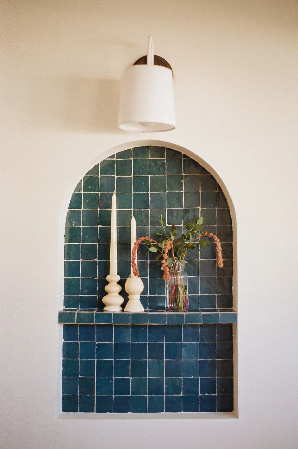 A custom arched tile niche using 2x2 mosaic zellige tiles in Aegean Sea tones. There is a mini shelf for souvenirs from travels. There is walnut sconce above with a linen shade.
