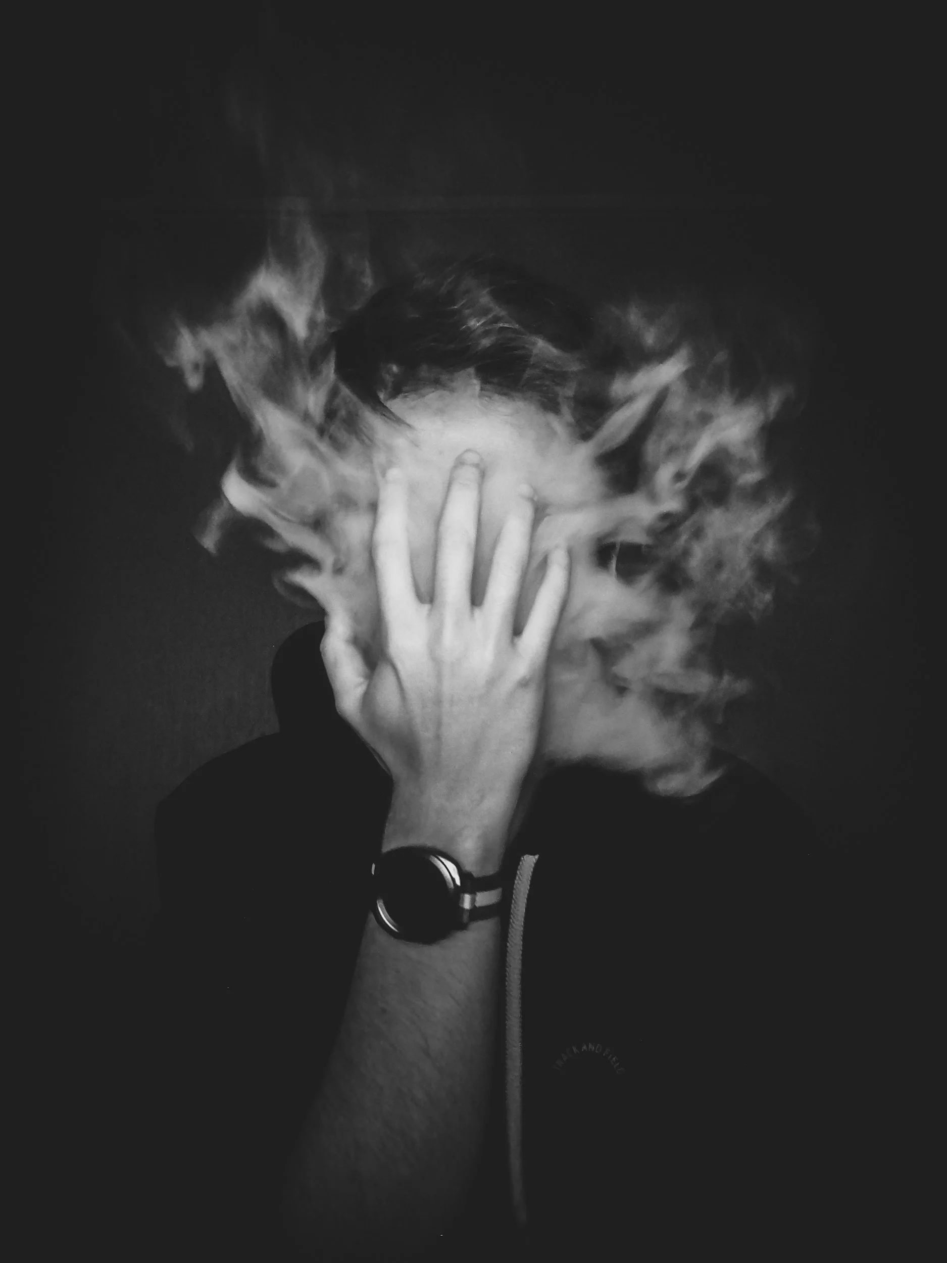 A person with a hand over their face, obscuring their features, surrounded by swirling smoke or vapor, black and white photo.
