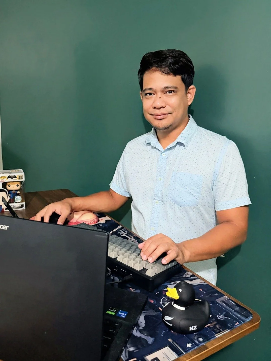 A man sitting at a desk with a computer, a duck-shaped rubber toy, and a figure of Superman, against a green wall.