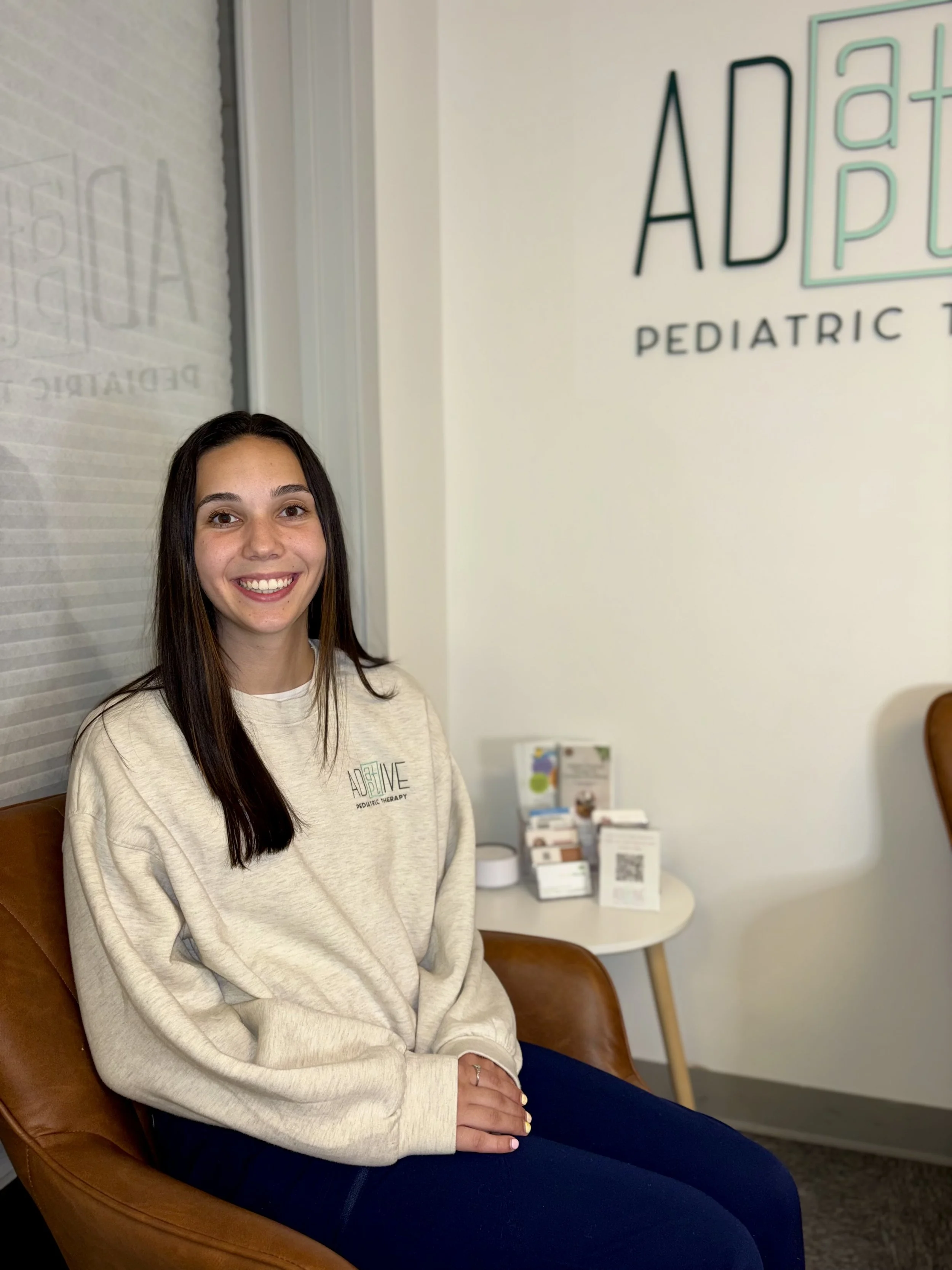 A smiling young woman with long brown hair, wearing a beige sweatshirt and dark pants, sitting on a brown leather chair in a medical office or waiting room. The background includes a white wall with a sign that says 'ADLVE PEDIATRIC' and some brochures or literature on a small round table.