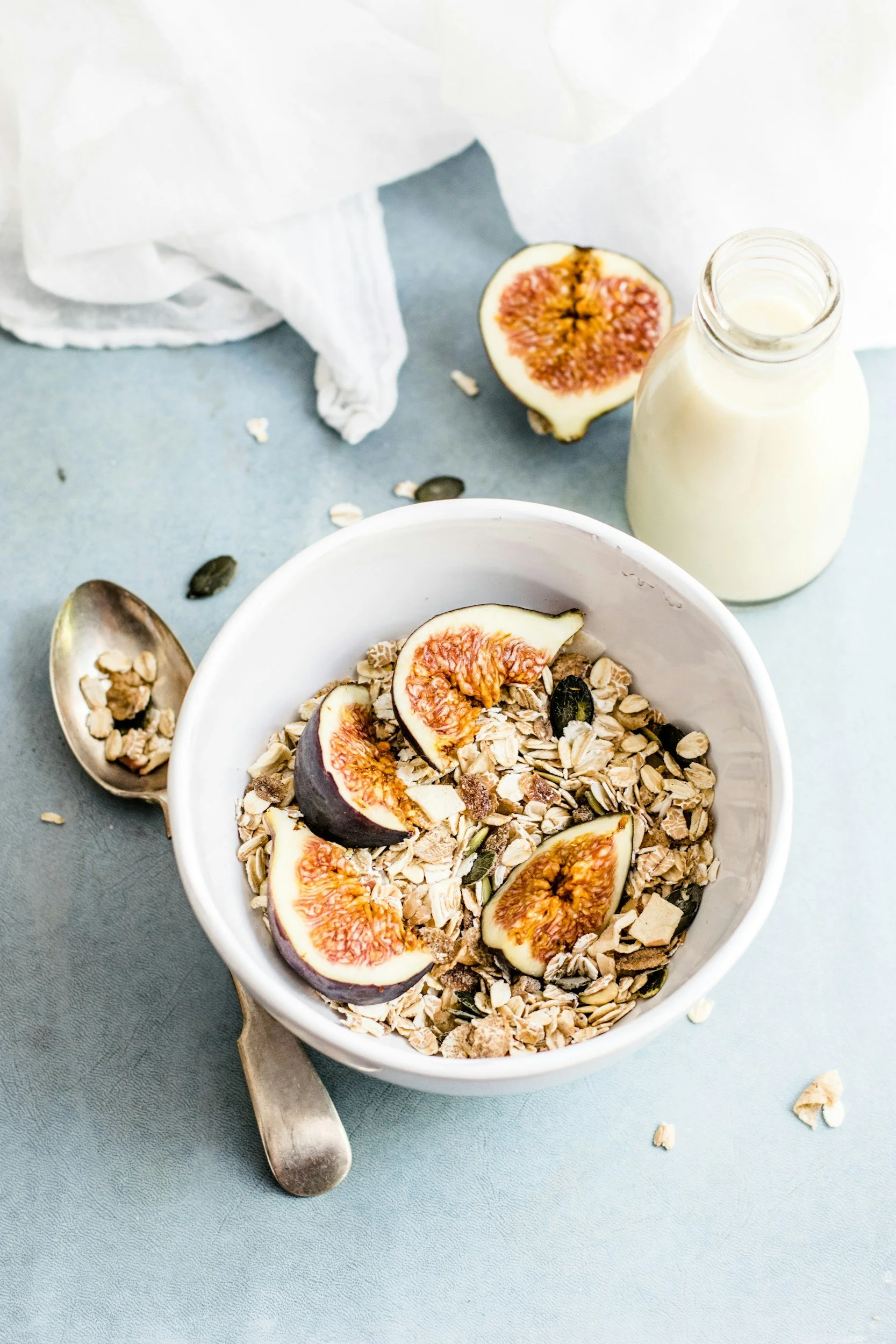 A bowl of granola with sliced figs on top, a bottle of milk, and a teaspoon on a light blue surface with scattered oats and fig pieces.
