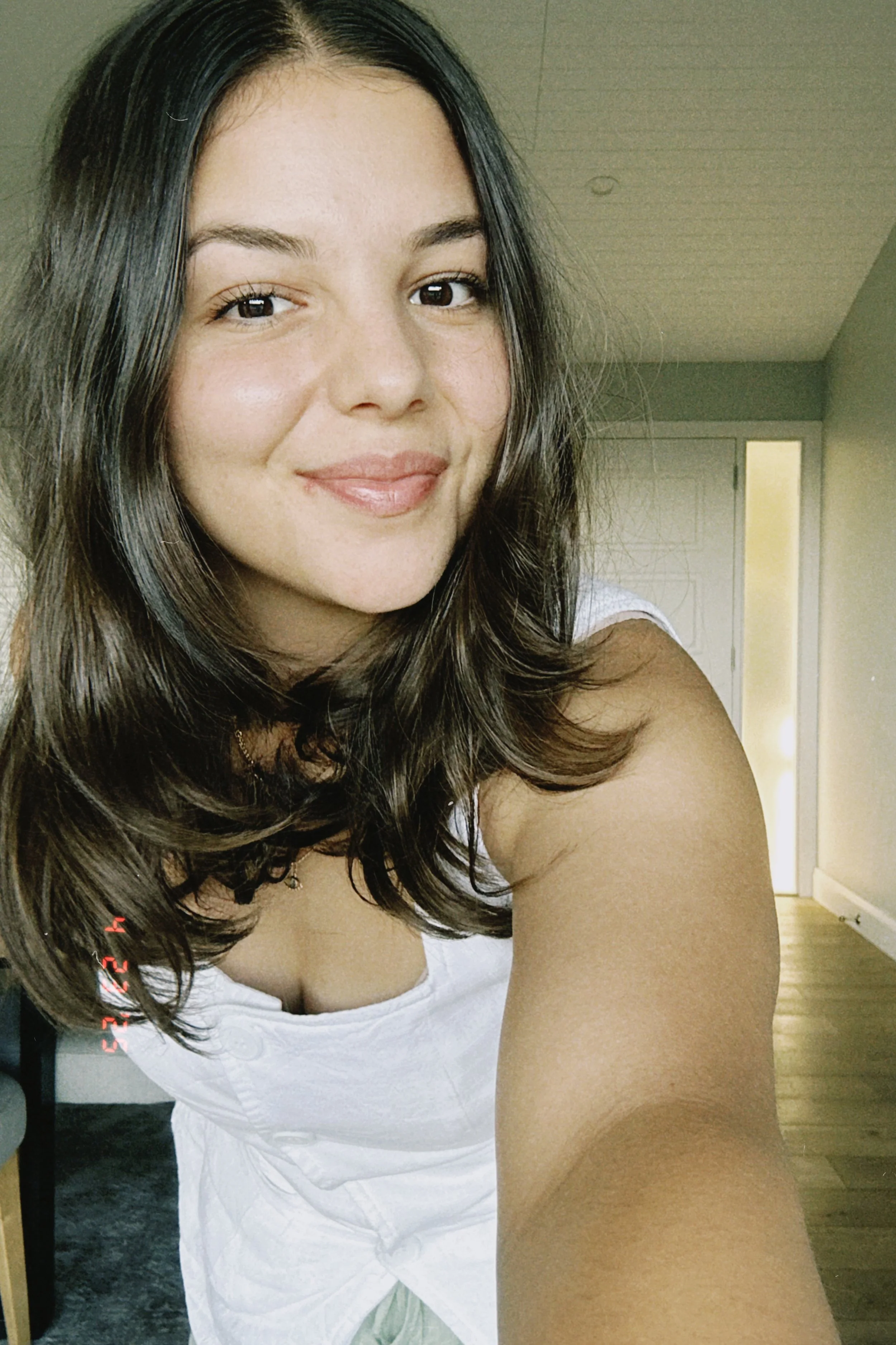 Close-up of a young woman with long brown hair smiling at the camera inside a room.