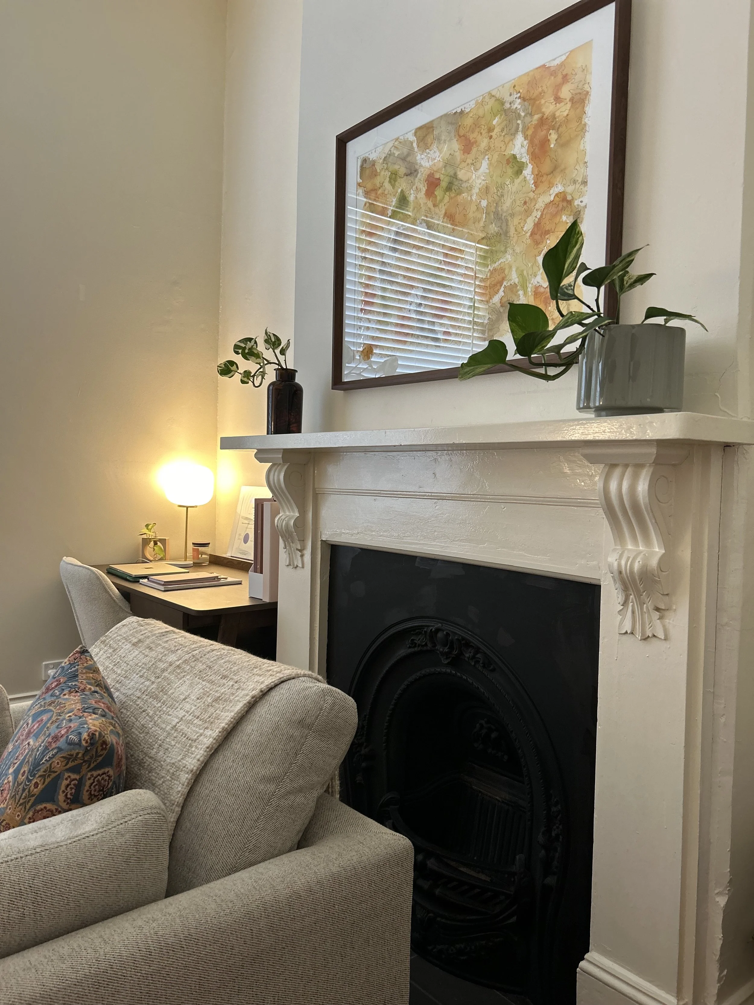 Living room with a beige couch, a white fireplace mantel with decorative carvings, a framed map of Paris on the wall above the mantel, a grey vase with green plant on the mantel, and a side table with a lamp, papers, and a small plant in the background.