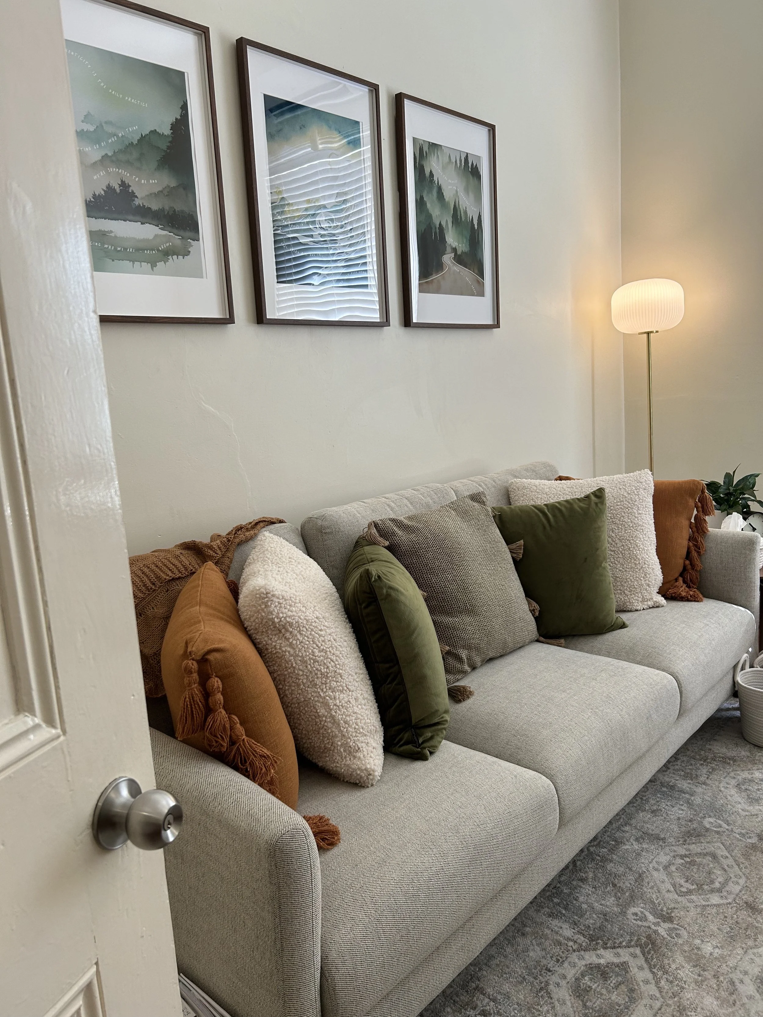 A beige couch with several throw pillows in white, green, and brown, in a living room. Behind the couch are three framed landscape prints on a cream wall, and a floor lamp with a white shade stands in the corner. A plant and some baskets are visible nearby.