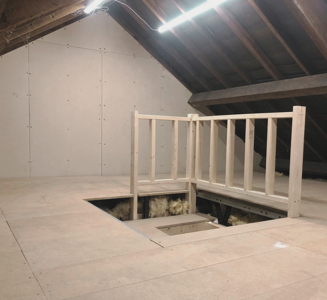 An attic room under construction with exposed wooden framing, insulation, and plywood flooring. The room has a sloped ceiling with visible roof beams and an opening in the floor.