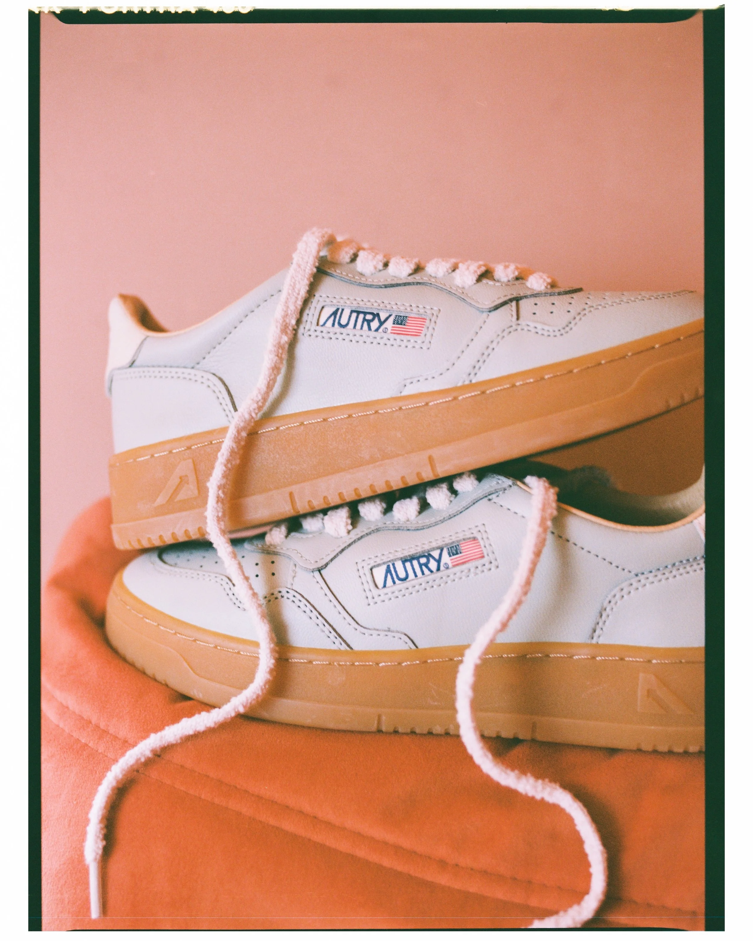 Close-up of a white sneaker with a gum sole, labeled 'AURRY,' placed on a peach-colored surface against a peach background, with a white shoelace in the foreground.