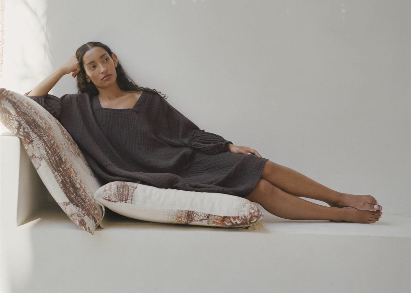 A young woman with dark hair lying on a white couch, propped up on her side with her head resting on her hand, wearing a loose black dress.