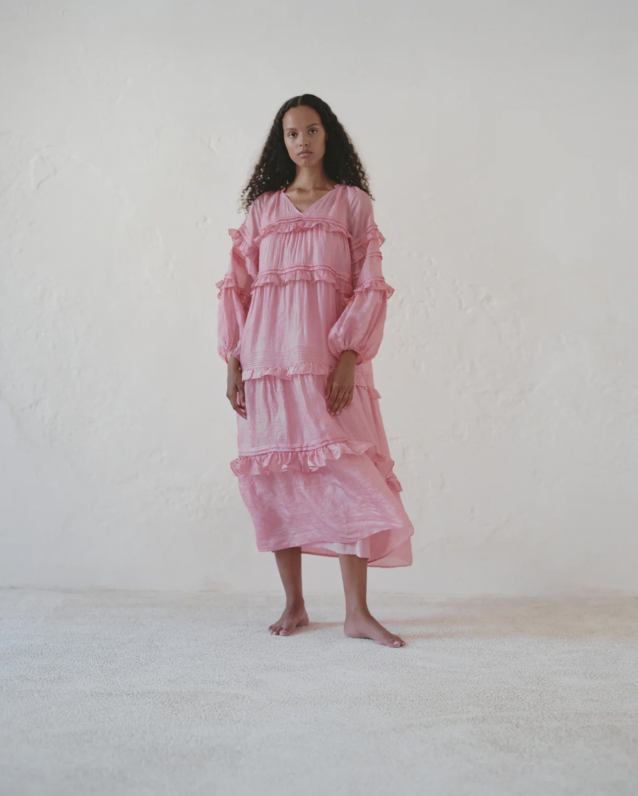 A woman wearing a long, pink ruffled dress standing barefoot on a light-colored carpet against a plain white wall.
