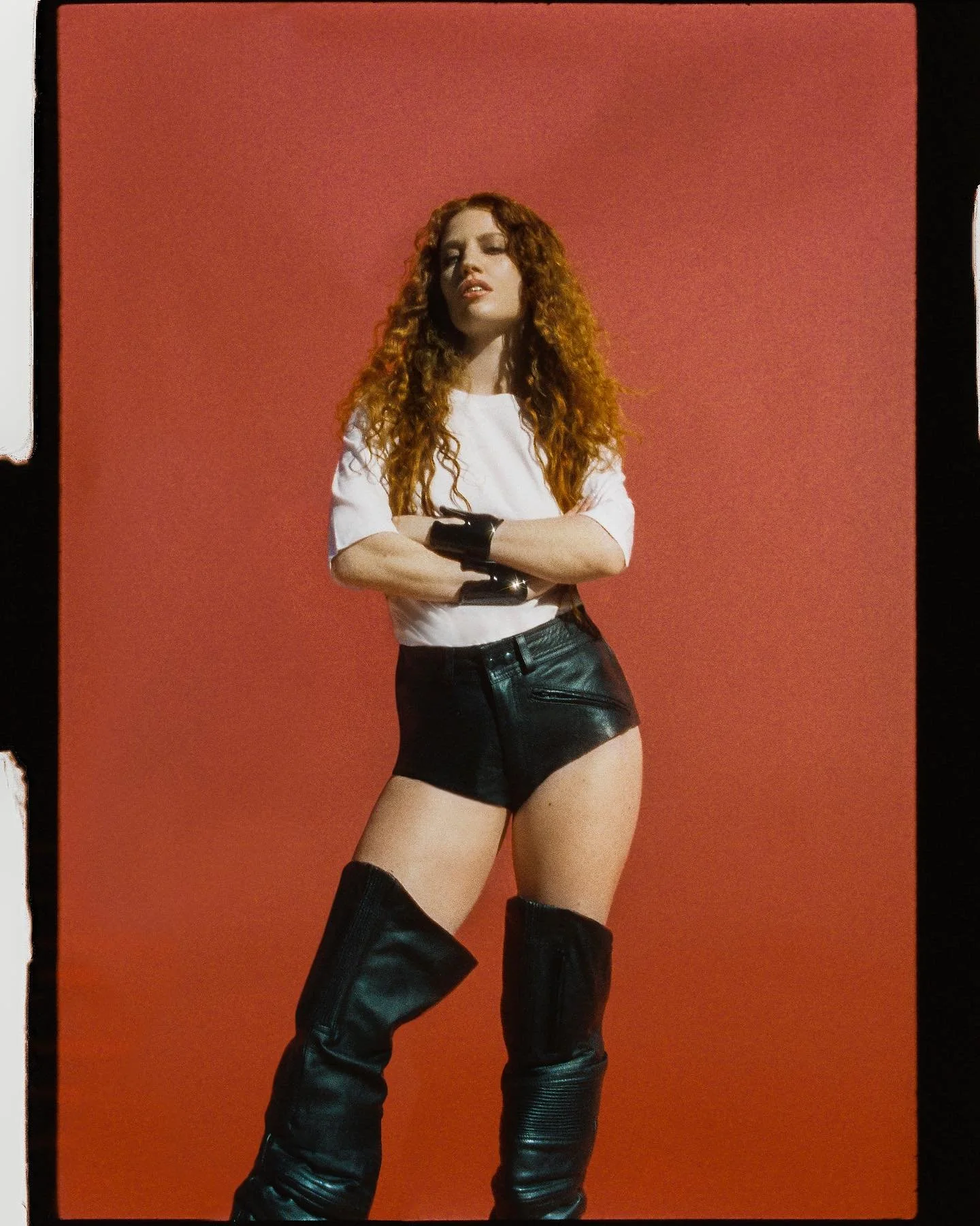 A woman with long, curly red hair standing against a red background, wearing a white T-shirt, black shorts, and tall black leather boots.