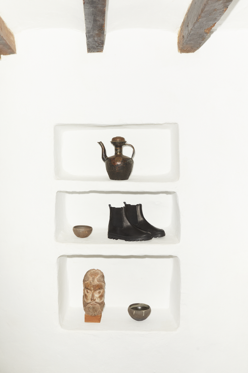 Built-in wall shelves with decorative items including an old ceramic teapot on the top shelf, a small bowl and black boots on the middle shelf, a carved stone face sculpture and a small bowl on the bottom shelf, set against a white wall with exposed 