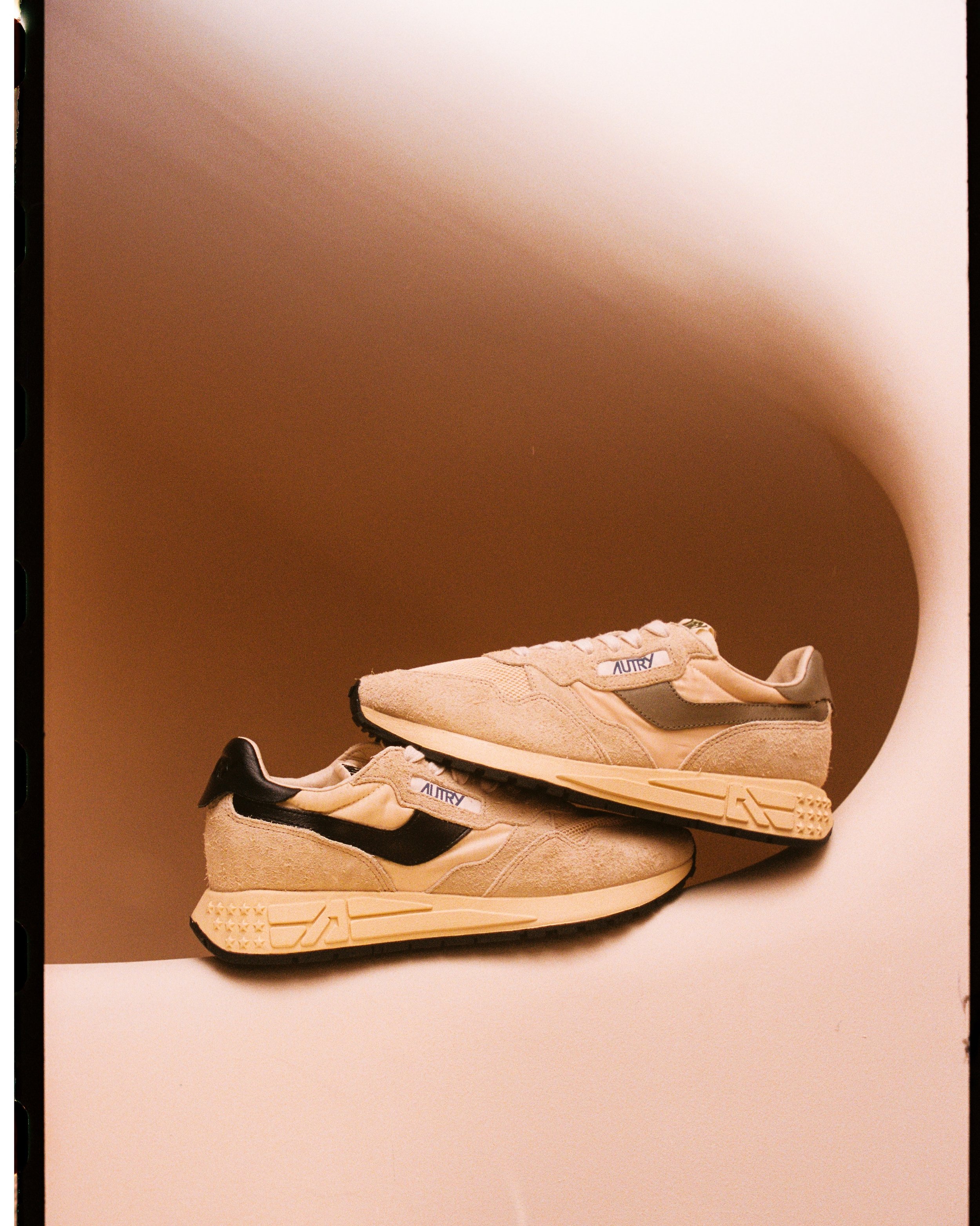 Pair of beige sneakers with black accents and 'AUTRY' branding, displayed on a beige platform against a plain beige background.