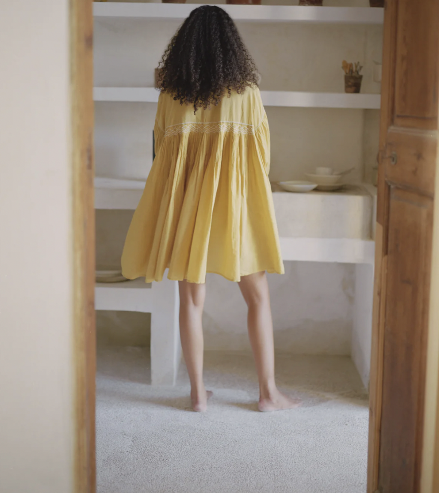 Person with curly hair wearing a yellow dress standing barefoot in a kitchen, viewed from behind.