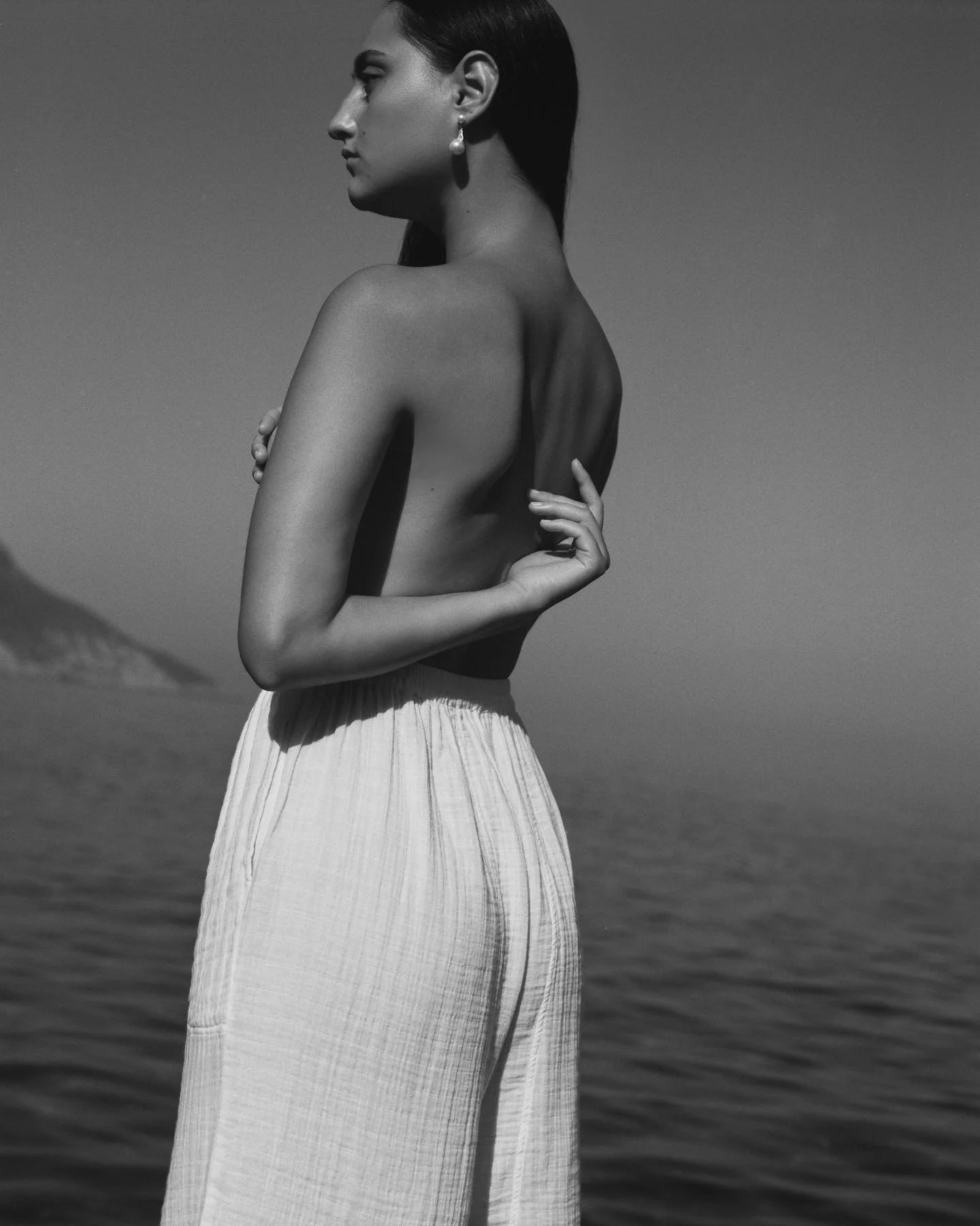 A woman with dark hair, wearing pearl earrings, is topless and standing near water with her arms crossed over her chest, looking to her left.
