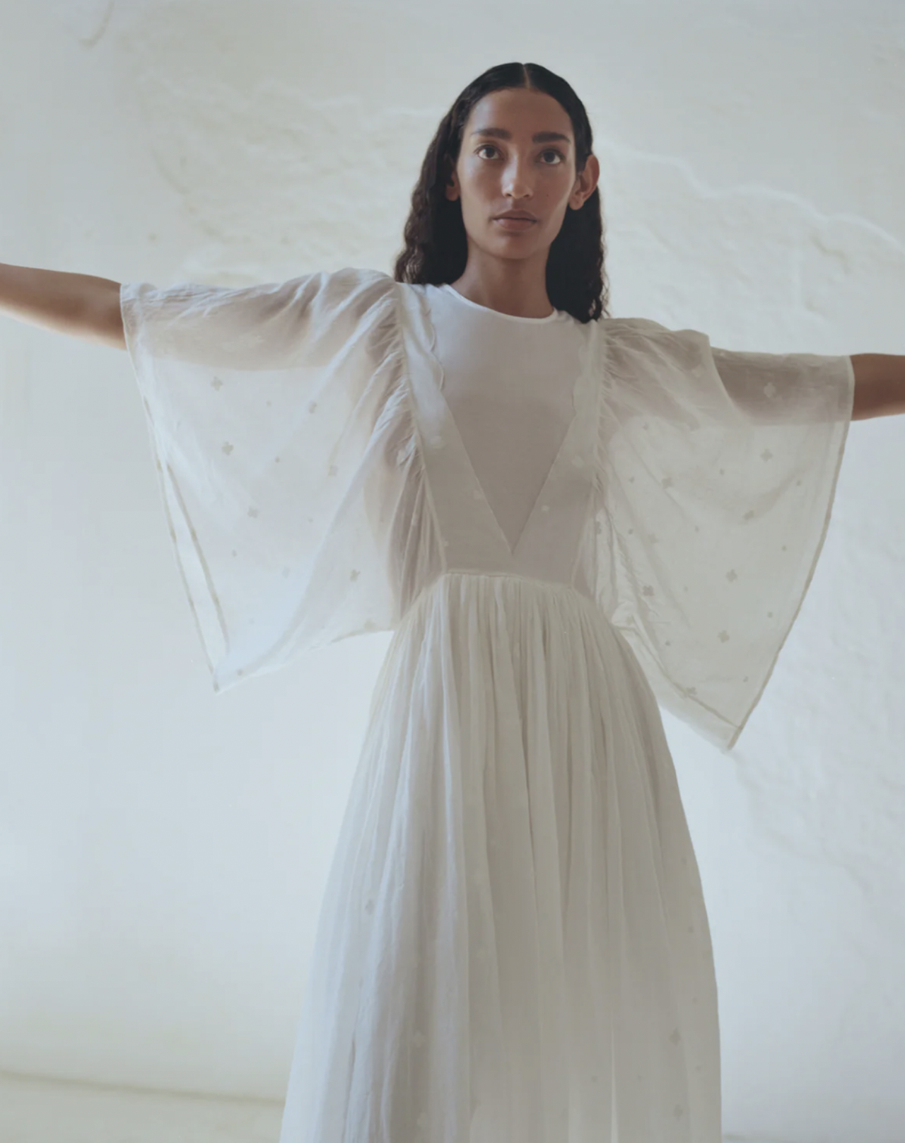 A woman with long dark wavy hair wearing a flowing white dress with sheer, embroidered sleeves, standing with arms outstretched against a light, textured background.