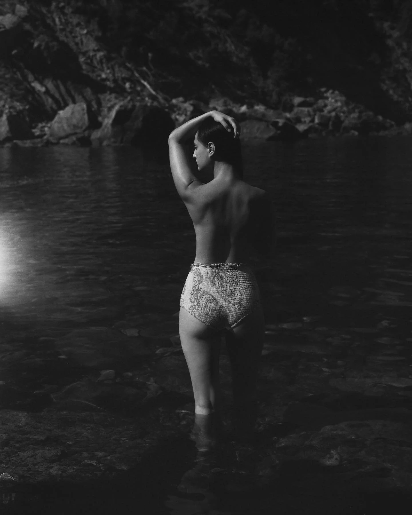 A woman standing in water at night, with one arm raised over her head, wearing patterned shorts, facing to the side, against a rocky background.