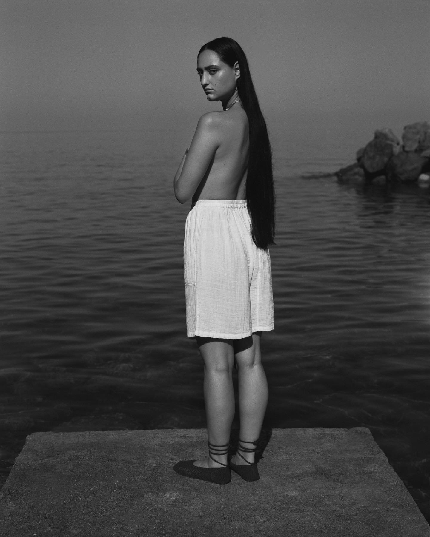 Black-and-white photo of a woman with long dark hair, standing on a concrete surface near water, looking over her shoulder at the camera, wearing loose light-colored shorts and sandals, with no top.