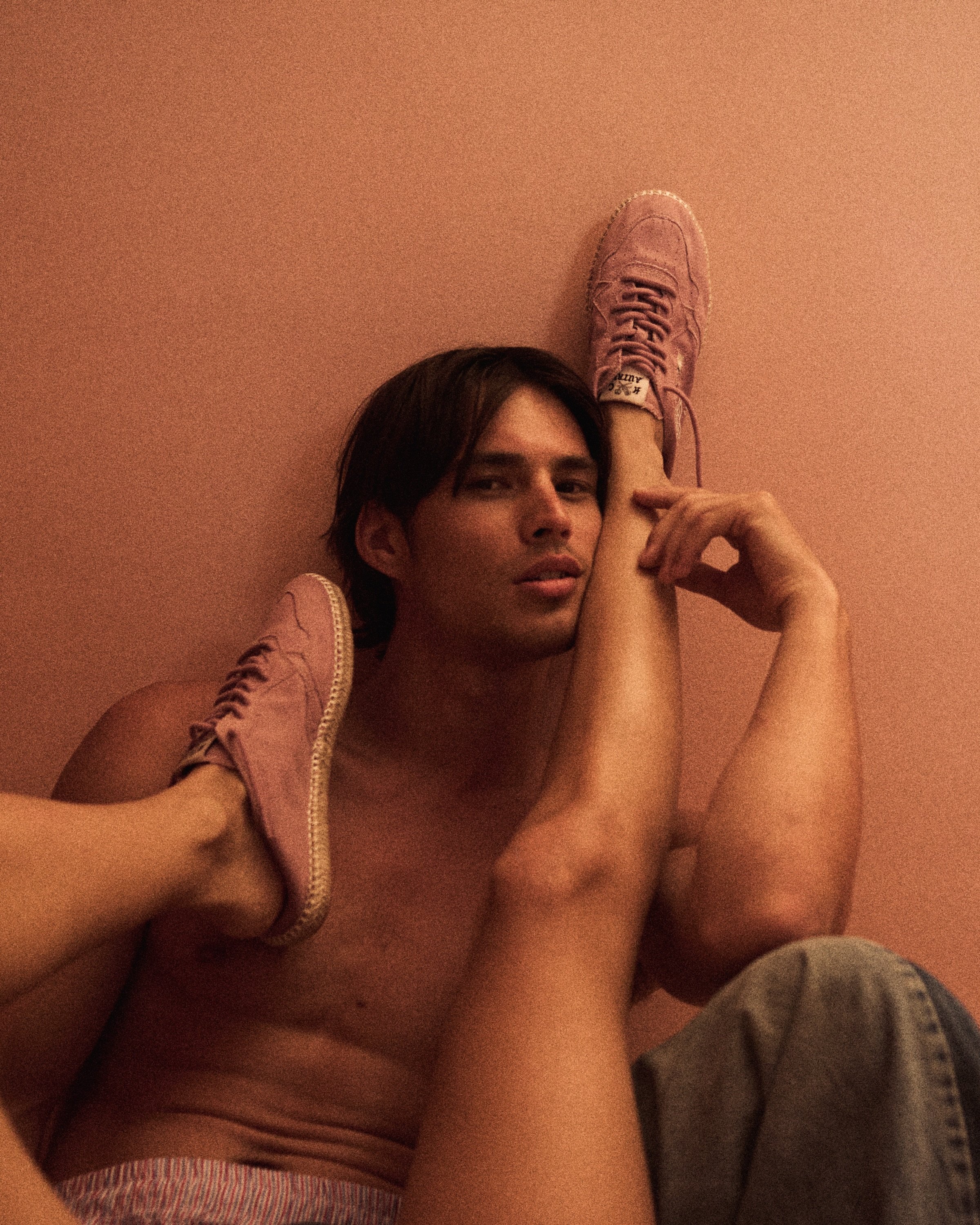 A person with light skin and dark hair, sitting on a pink floor, looking at the camera while resting their right arm on their knee, wearing pink sneakers and striped shorts.