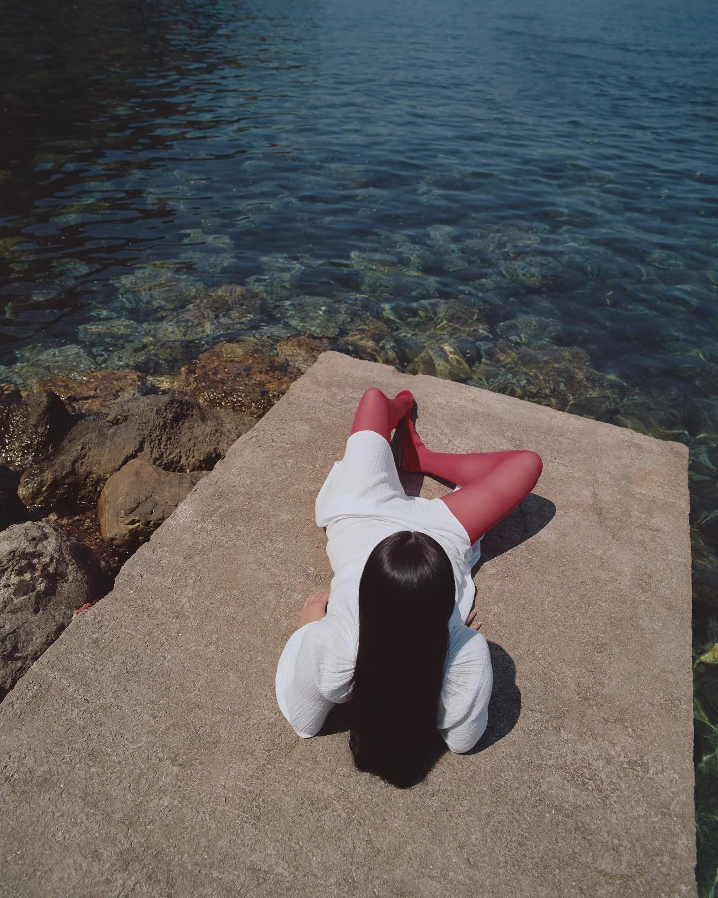 Woman with long dark hair, dressed in a white top, red tights, and red shoes, lying on a concrete surface by the water with rocks visible nearby.