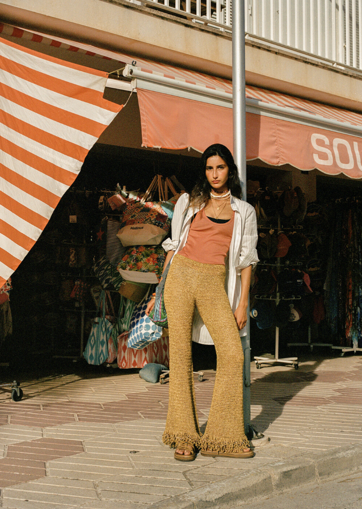 A woman standing on a city sidewalk in front of a street stall with striped awning selling bags and clothing. She is wearing a light-colored shirt over a peach top, patterned wide-leg pants, and sandals, with a necklace and a crossbody bag.