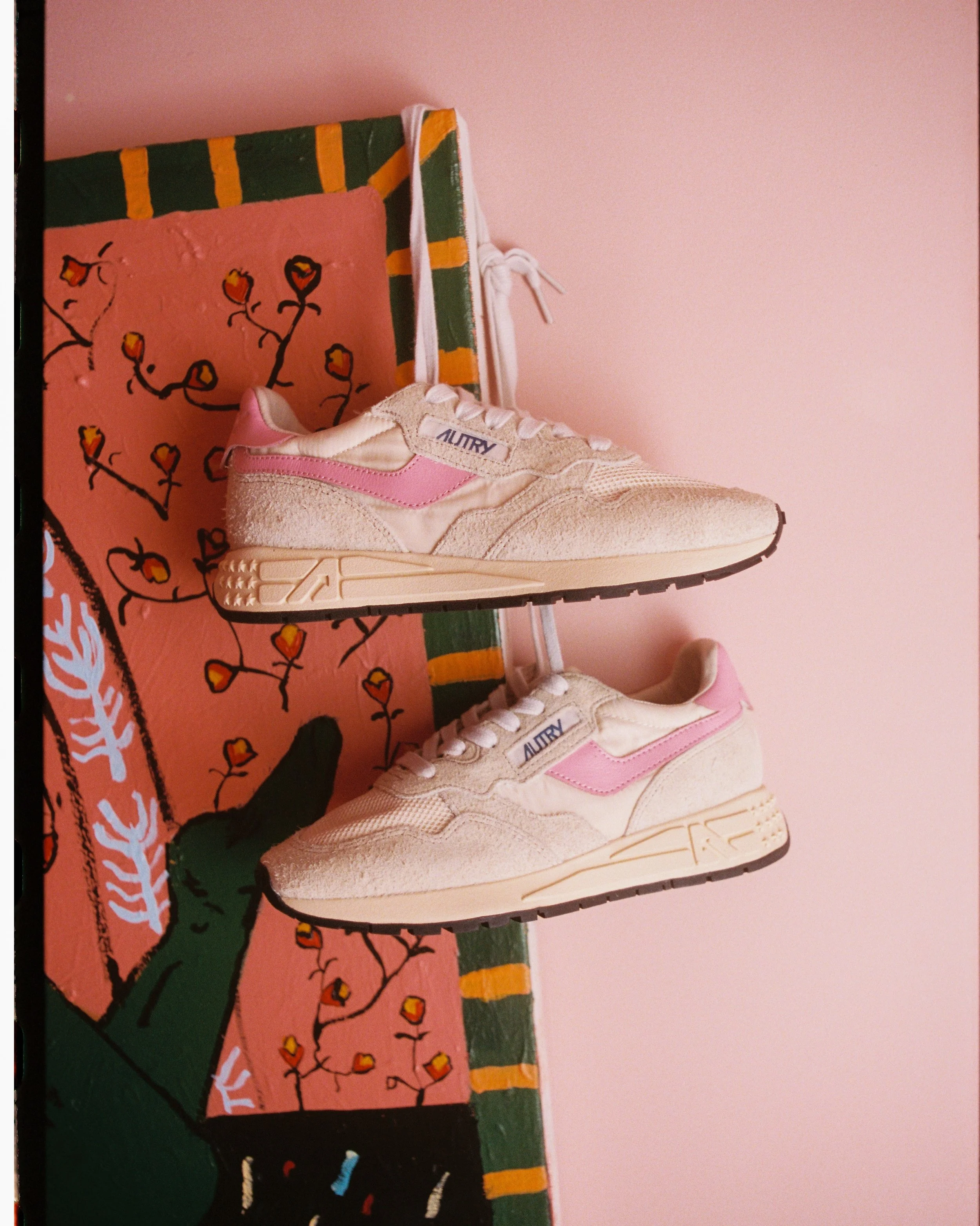 A pair of beige sneakers with pink accents placed on a decorative rug, with a pink wall in the background.